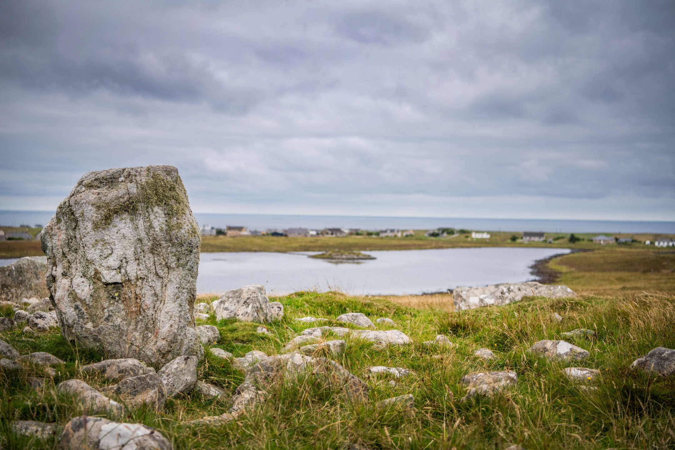 Single standing stone with loch and houses in background under cloudy sky.
