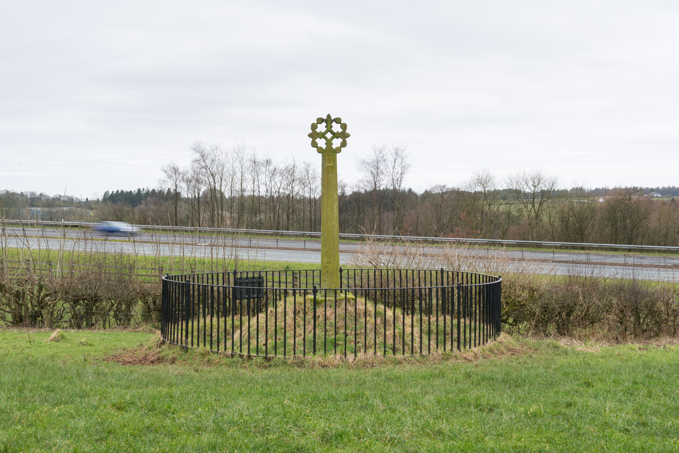 Stone cross monument surrounded by a black fence in a grassy area by a road