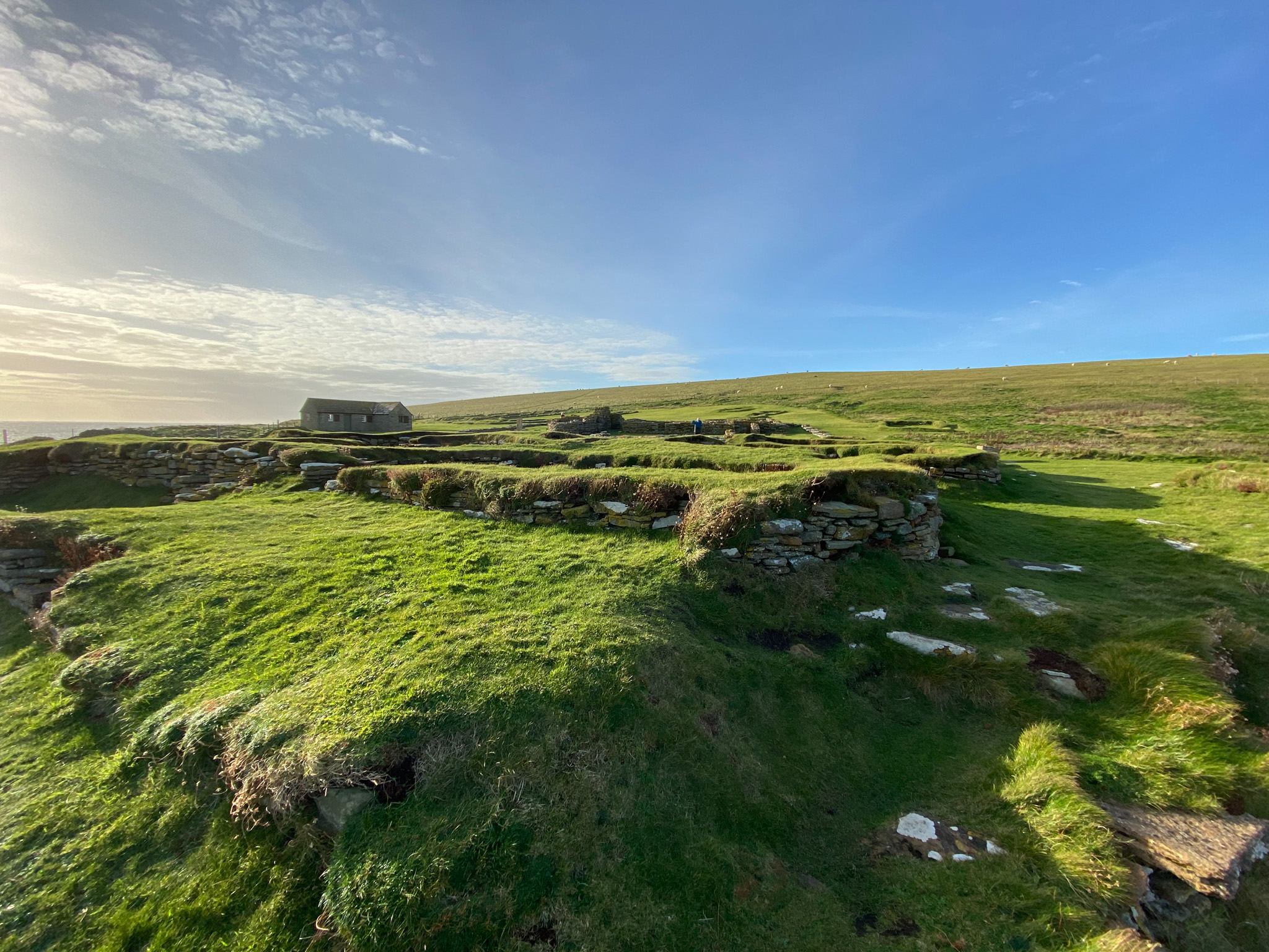 Ruins of ancient stone structures covered in grass, set on sloping green land with a small stone building in the distance.