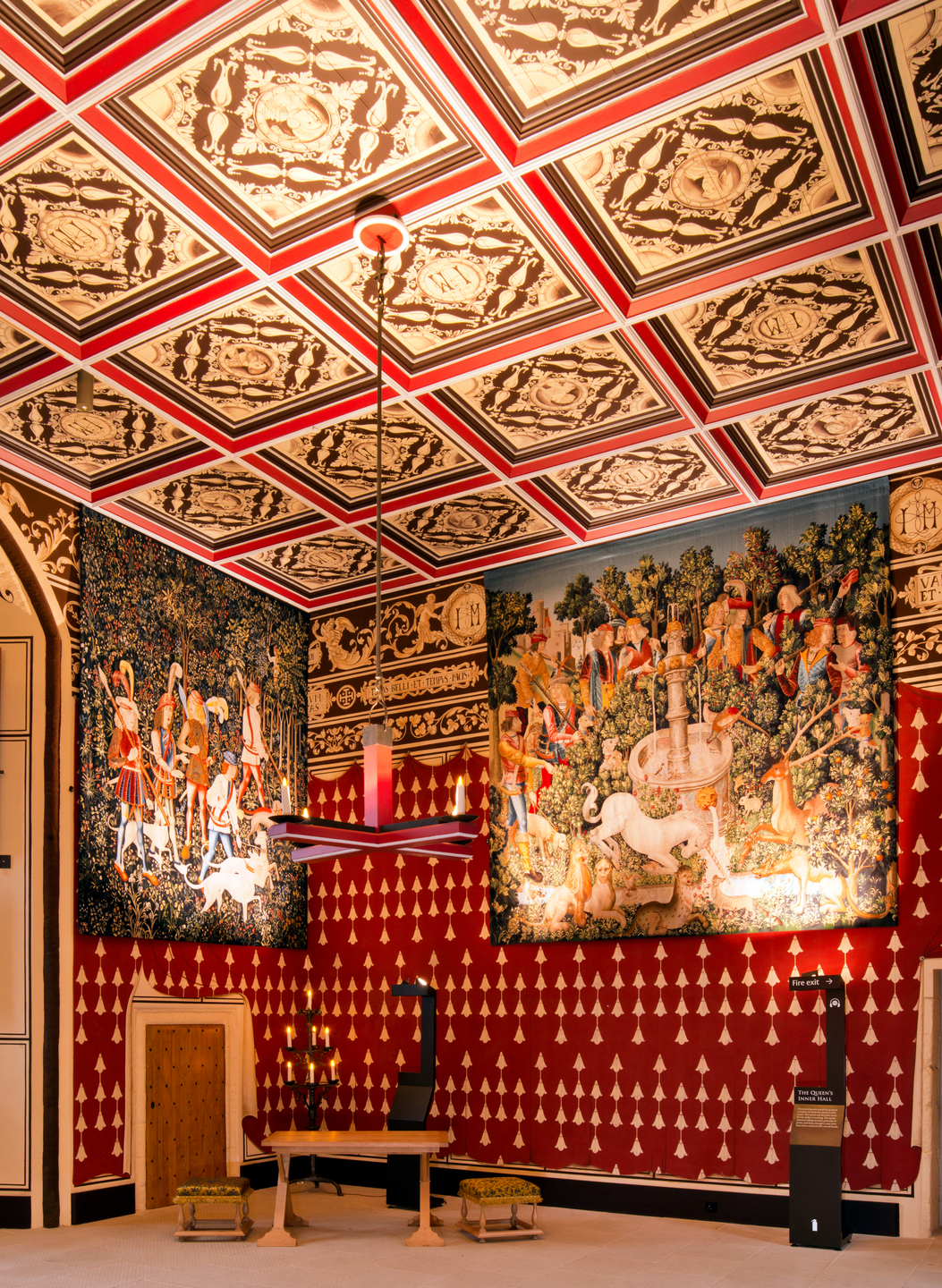 A formally decorated, medieval style interior of a room with high ceilings. There are two large tapestries on the wall, with people appearing on them. The walls and ceiling are red with gold detail. 