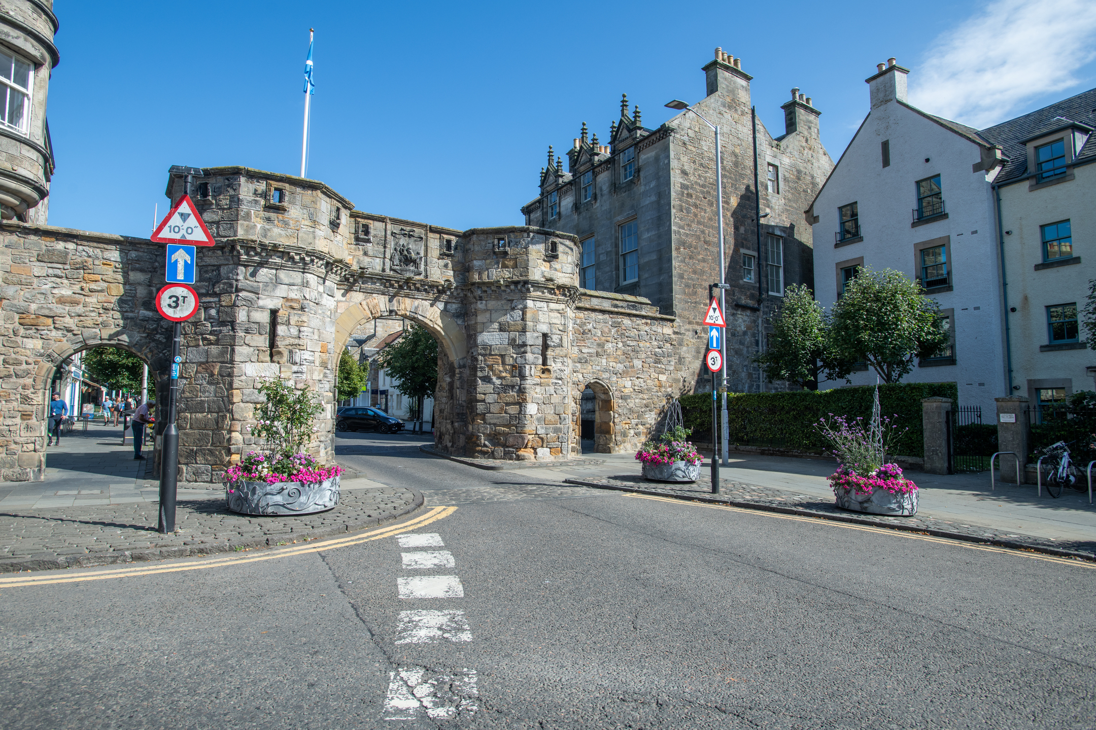 Angled street view showing the full West Port gateway with its central arch and adjoining smaller pedestrian arch. The structure is built from rough-cut stones in varying tones. To the right stand tall historic buildings with crow-stepped gables. Flower planters and traffic signs line the foreground