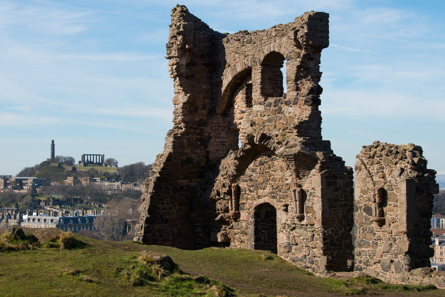 Ruins of St Anthony’s Chapel with Calton Hill and its monuments visible in the background under a clear sky.