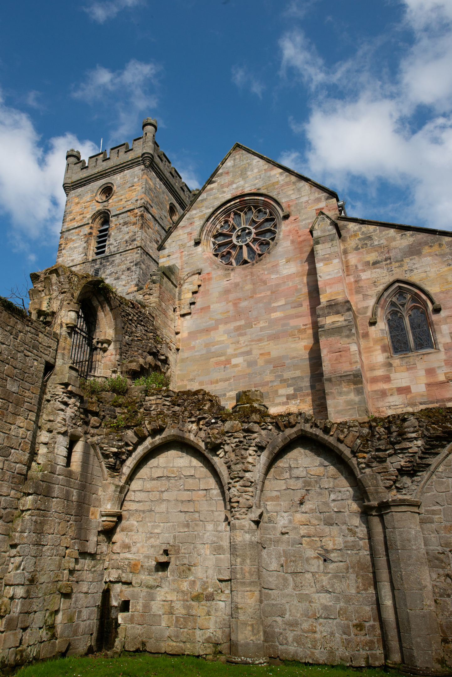 Culross Abbey’s tower and rose window viewed from the ruins on a bright day, with blue sky overhead and the remains of arched stone walls in the foreground.