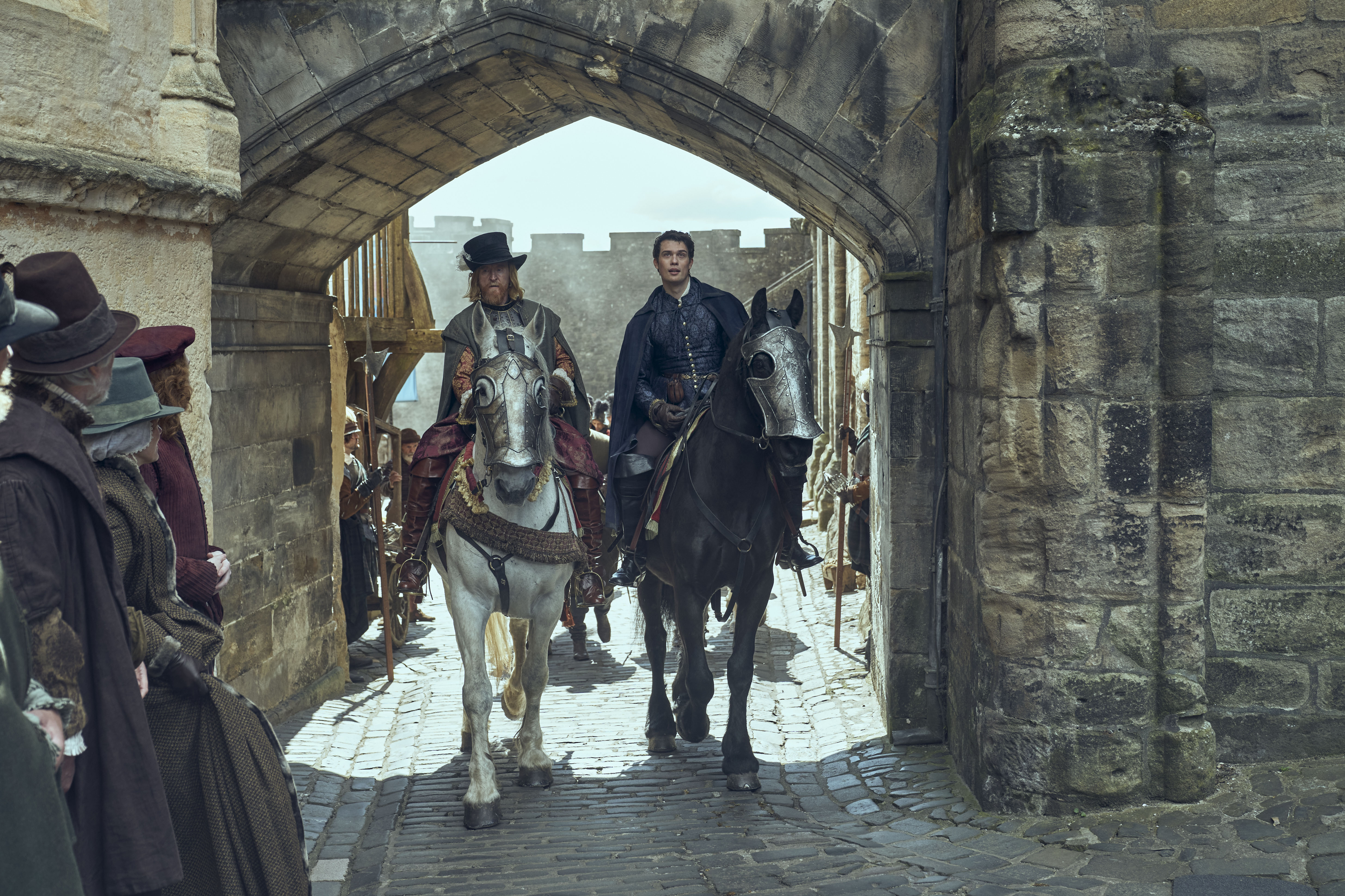 Two men on horseback ride through the stone gate of a castle. The lead rider on a white horse wears elaborate historical dress, while the rider on the dark horse wears a dark cloak. Townsfolk watch them pass.