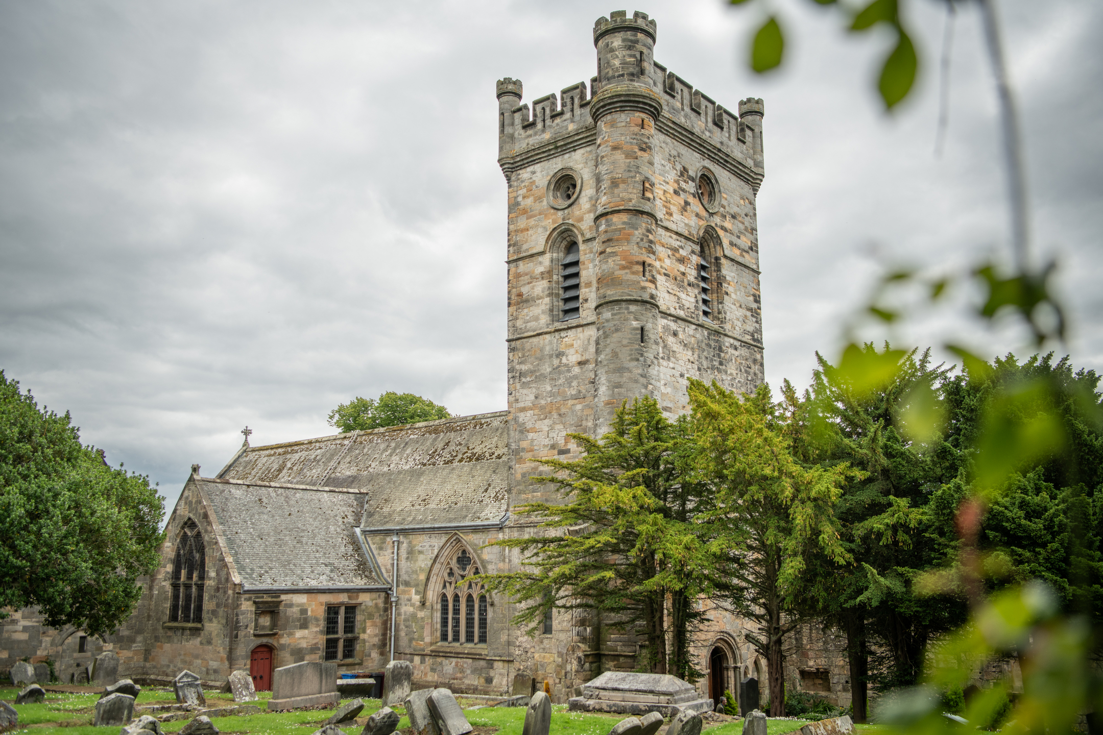 The exterior of Culross Abbey on an overcast day, showing the tall square tower, arched windows, and surrounding churchyard with scattered gravestones and mature trees.