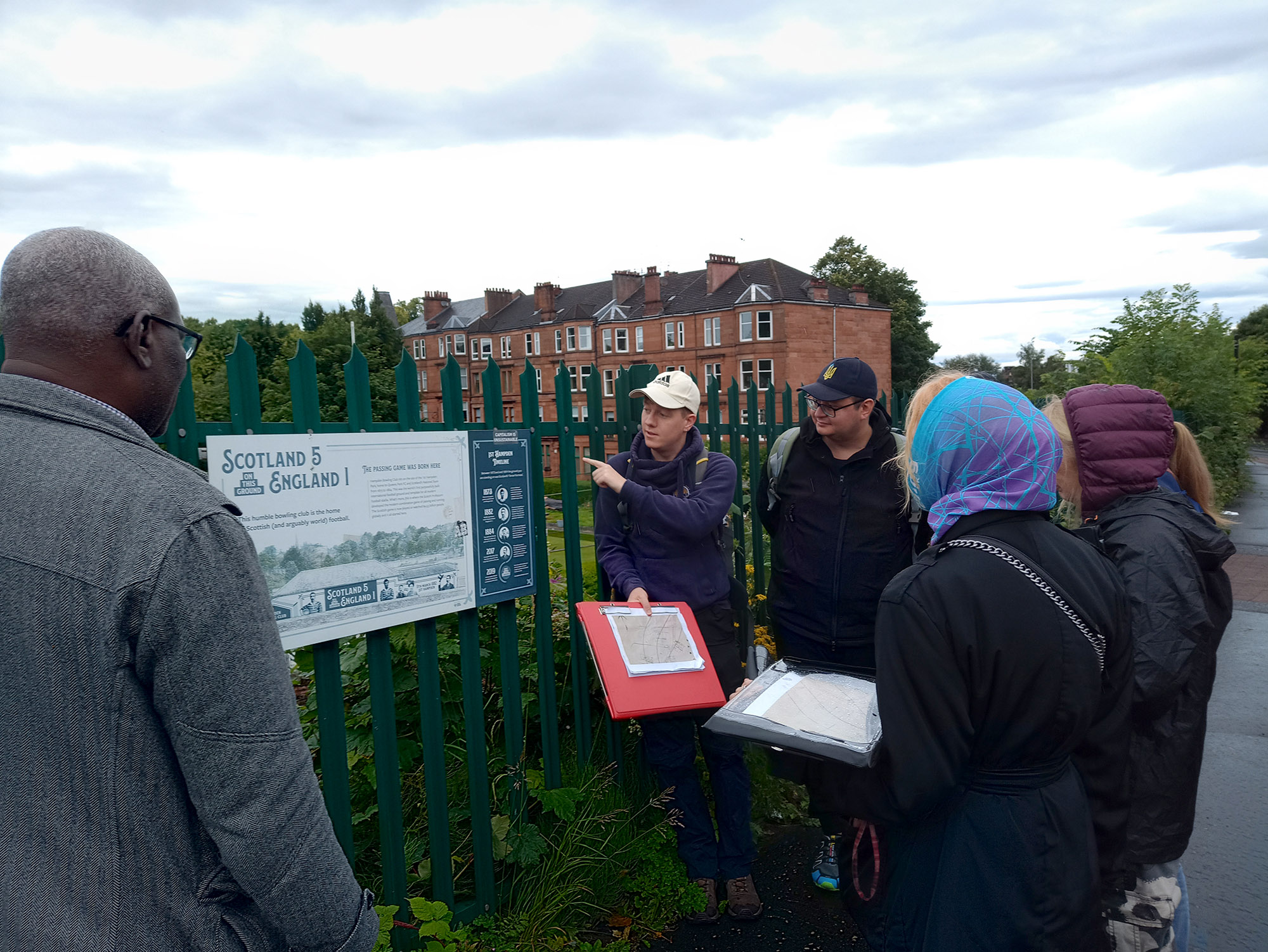 A group of people stand beside a green metal fence listening to a guide during a heritage walking tour. The guide points to an information board titled “Scotland 5 England 1” while holding a folder with maps and notes. Behind the group are red sandstone tenement buildings and trees under a cloudy sky. The scene shows participants learning about local history and archaeology in an urban outdoor setting.