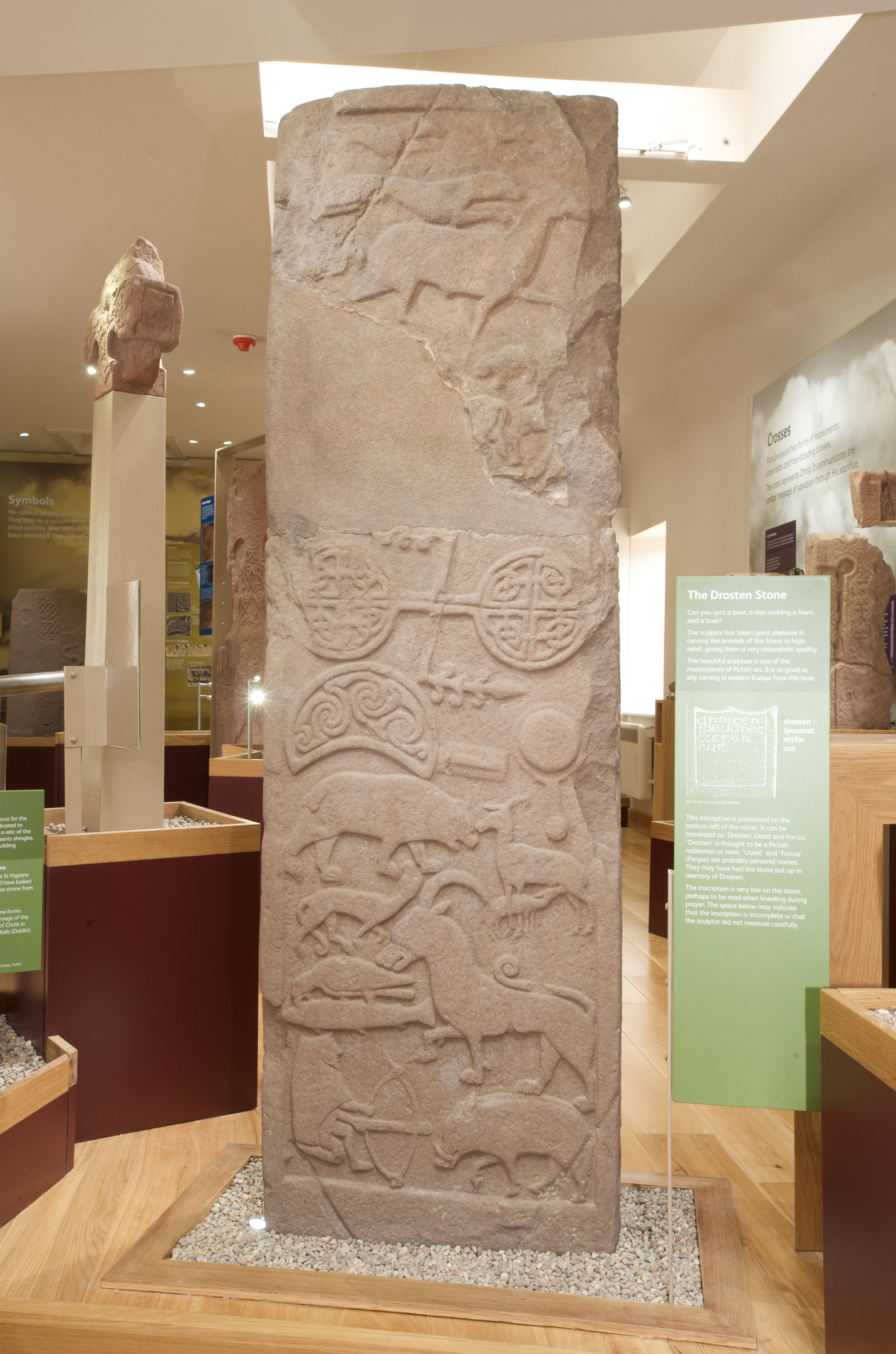 Full view of the Drosten Stone, a tall sculptured Pictish stone with intricate carvings of animals, interlace designs, and circular motifs, displayed upright with an interpretation panel beside it.