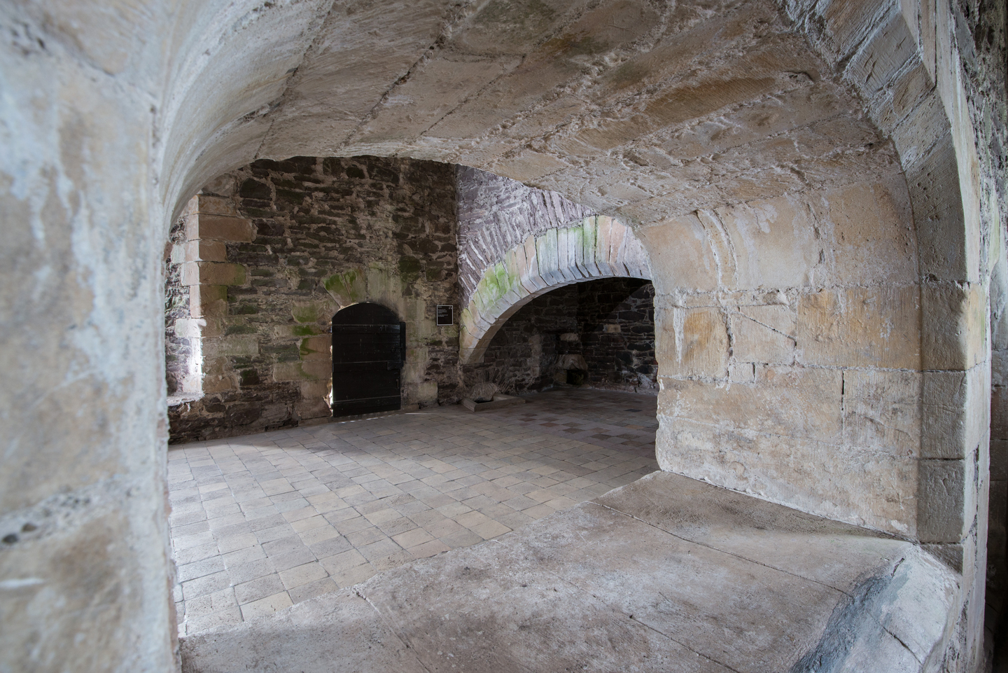 Interior of a historic stone room, through a small pale stone arch. There is a small but wide wood door on the wall opposite, as well as a broad arch. 