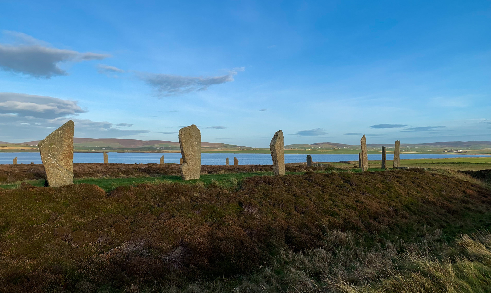 An enormous stone circle erected beside a loch. Conditions are tranquil, with a bright blue sky and still waters on the loch.