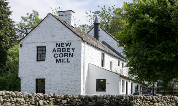A stone building painted white surrounded by trees with a low stone wall running in front of it. On the side of the building the words New Abbey Corn Mill are painted in black.