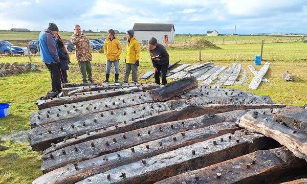 A group of people in rain macs, hats and boots stand in a green field. In front of them is a larger weathered structure of timber beams, with more wooden beams laid out behind them. There are stone cottages dotted along the landscape behind them and the sun is shining