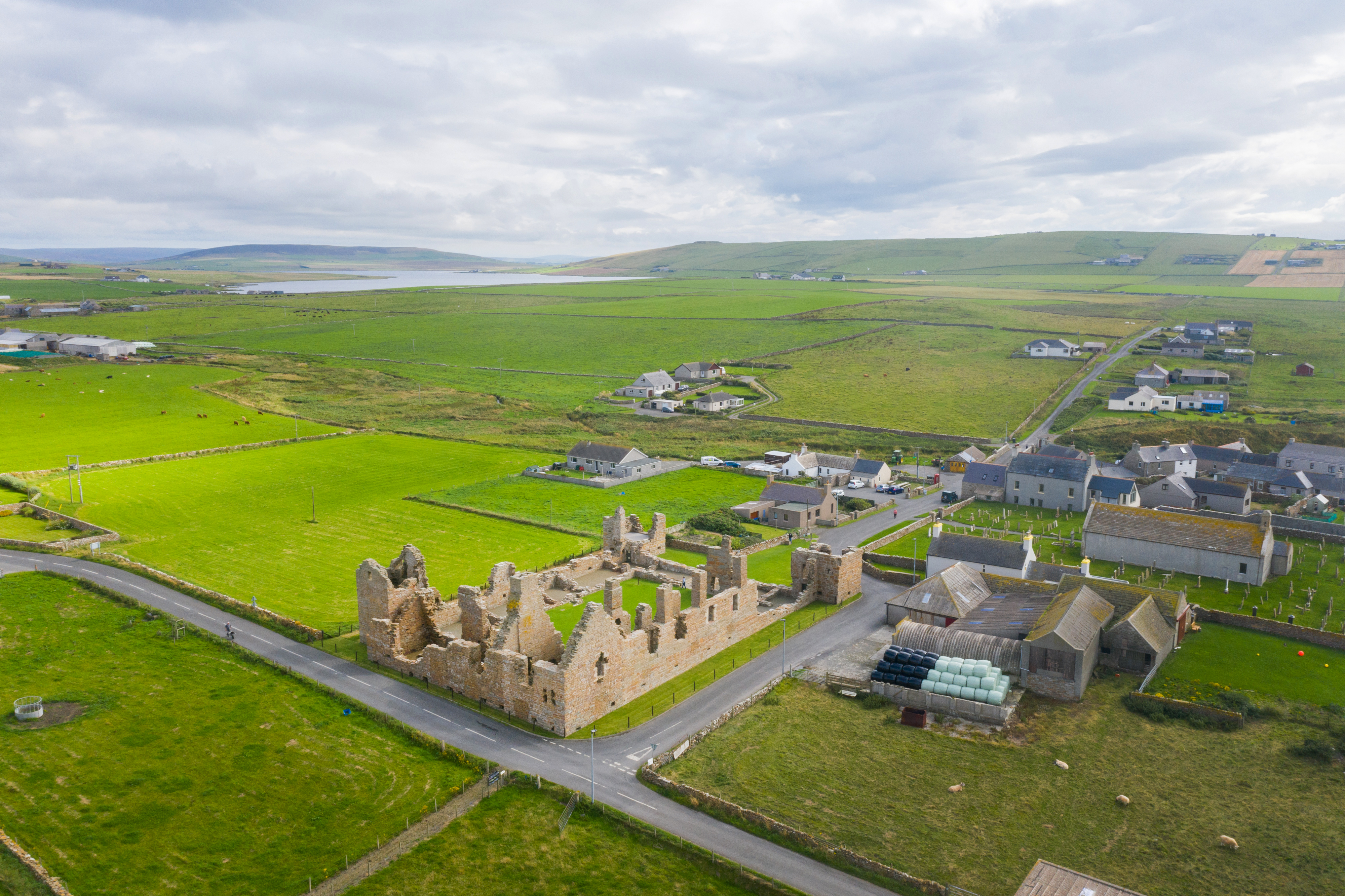 The palace sits among wide, open fields with scattered farm buildings and rolling hills in the distance. The layout of the village roads and stone boundaries is clearly visible from above.