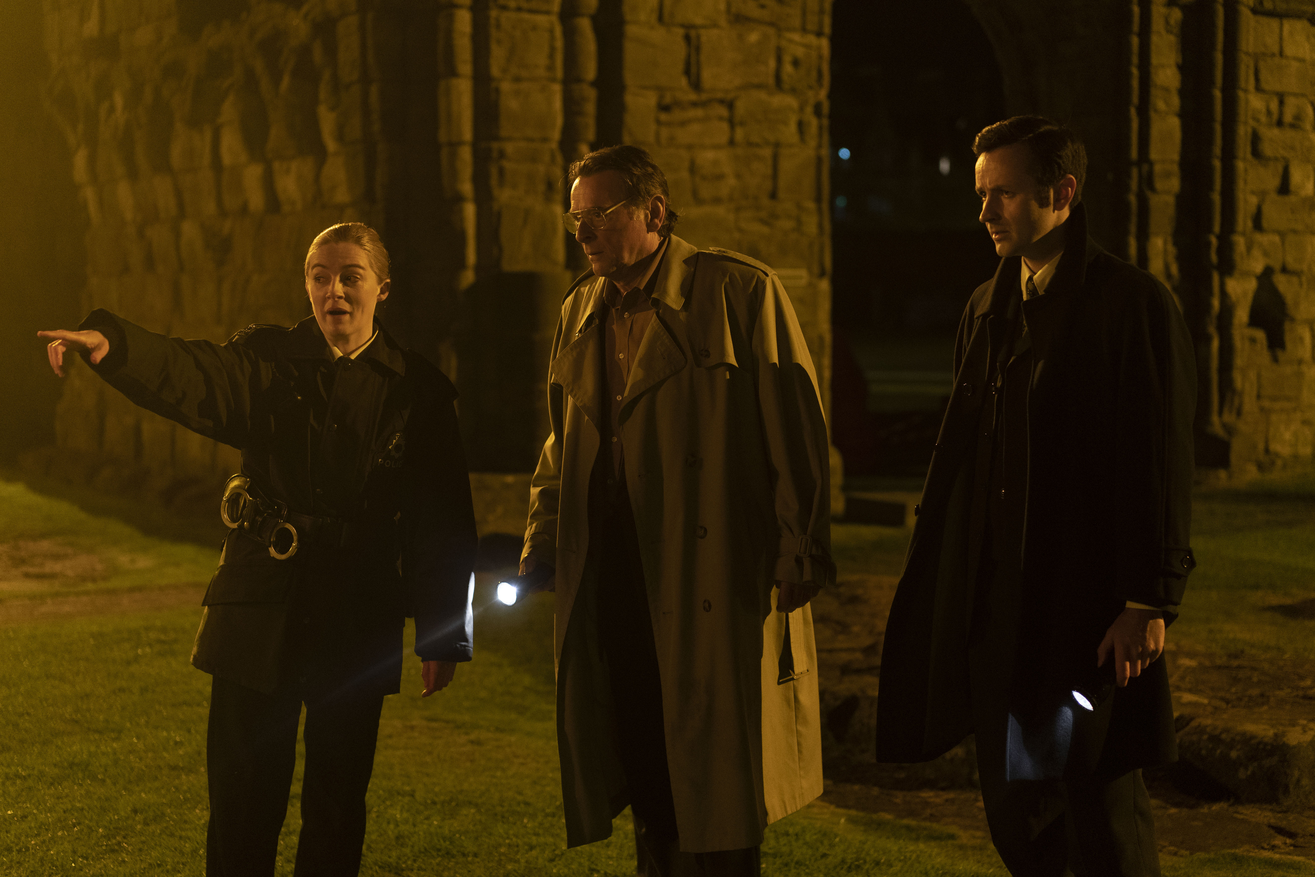 Three investigators stand outside ancient stone ruins at night. A young female officer points to something off‑frame while two men beside her, both holding torches, listen intently.
