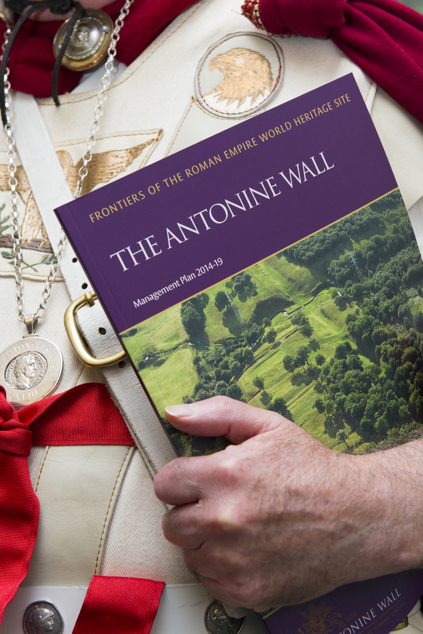 Close up image of a man wearing a Roman soldier costume holding a booklet which reads "The Antonine Wall Management Plan 2014 - 19"