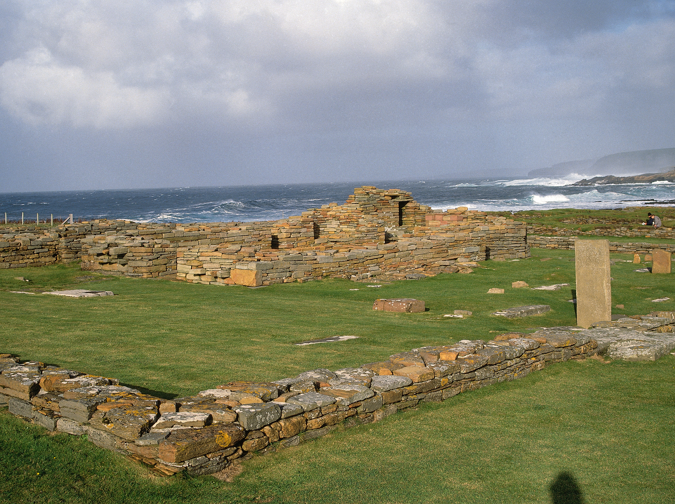 Ancient stone foundations on grassy ground near the edge of a rough, windswept coastline with waves crashing in the background.