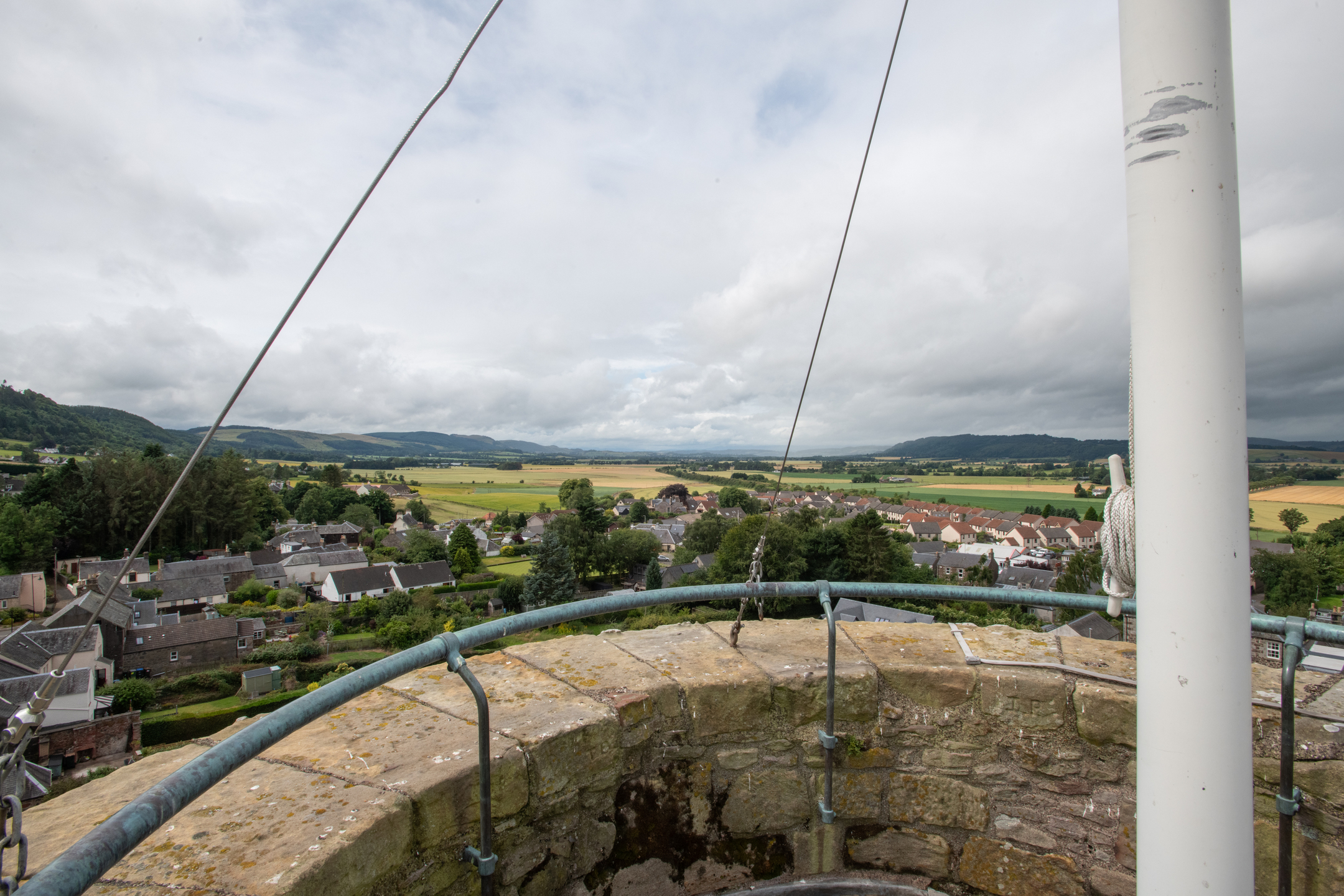 A panoramic view from the top of Abernethy Round Tower overlooking rooftops, fields, and rolling hills under a bright, cloudy sky, with the tower’s stone rim and flagpole in the foreground.