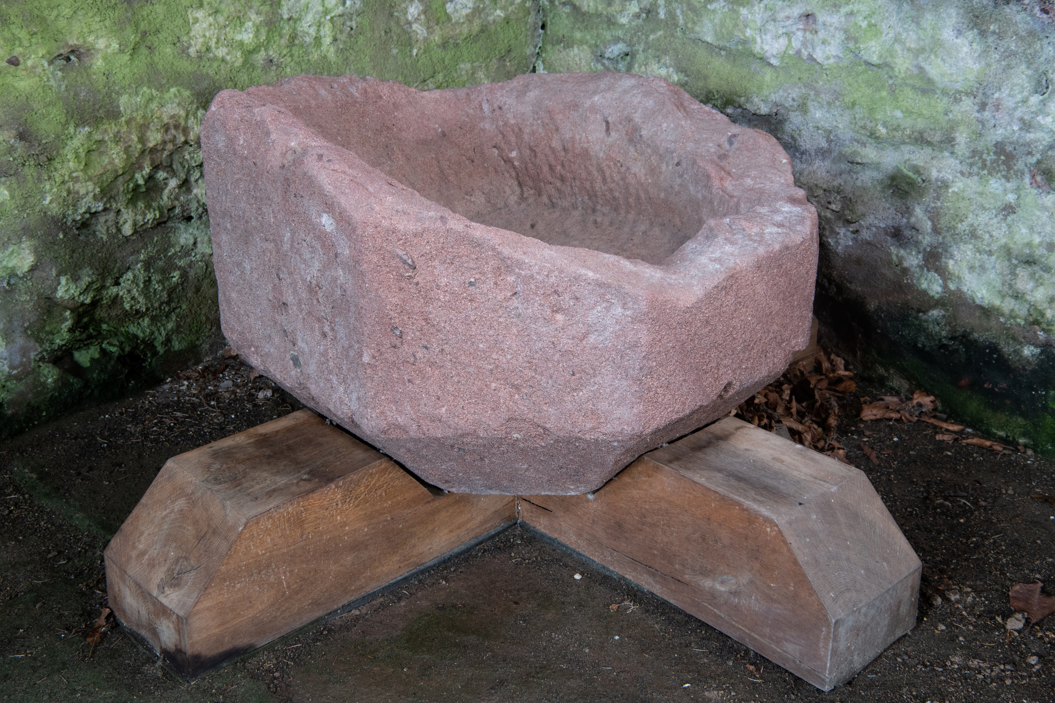 Rough-hewn red sandstone basin with a deep hollow interior, mounted on a sturdy wooden cross-shaped support. The basin sits on a stone floor against a moss-covered wall, suggesting historic use as a font or trough