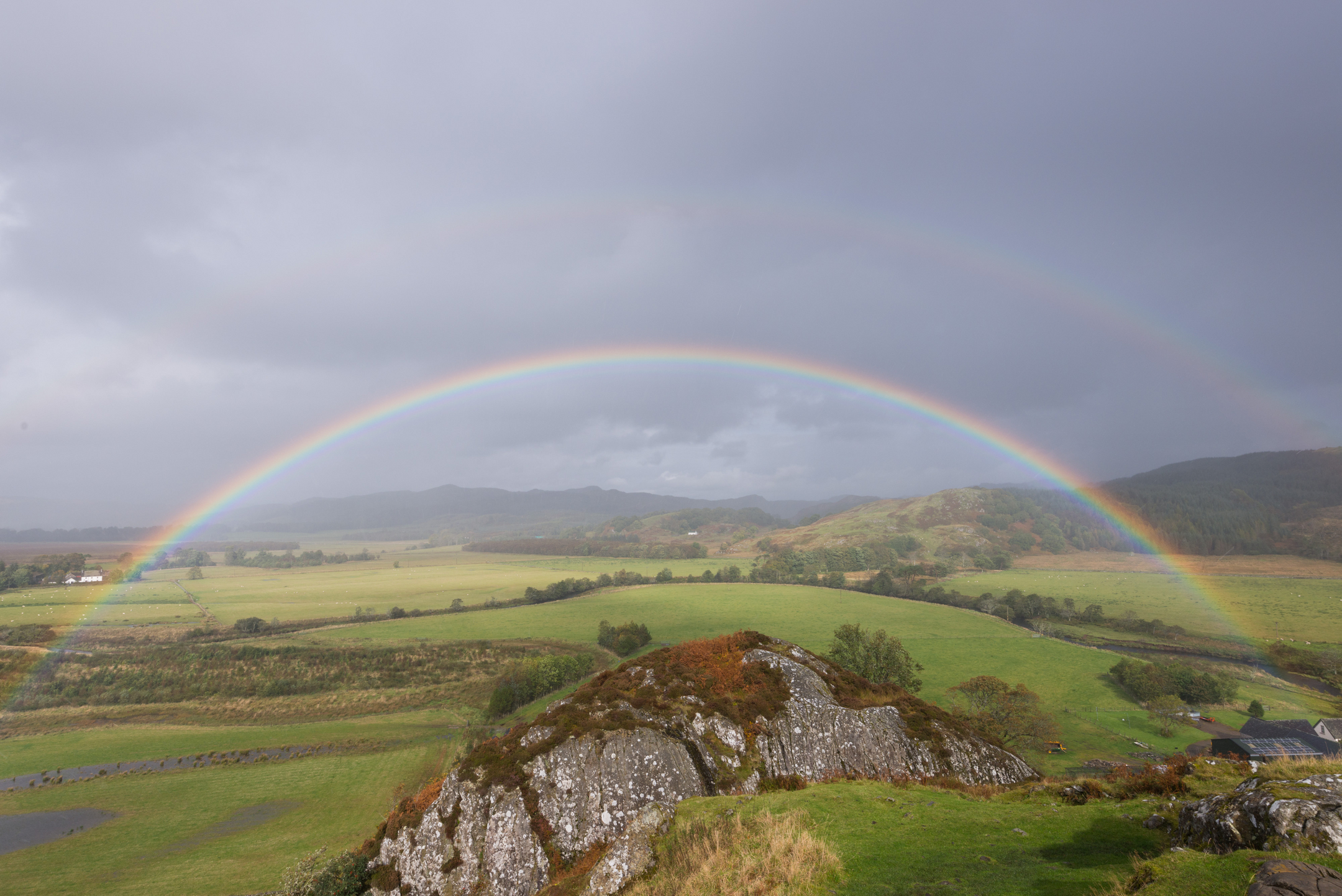 A bright rainbow arching over Dunadd Hill and the surrounding green valley, with a second faint rainbow visible against a grey sky.