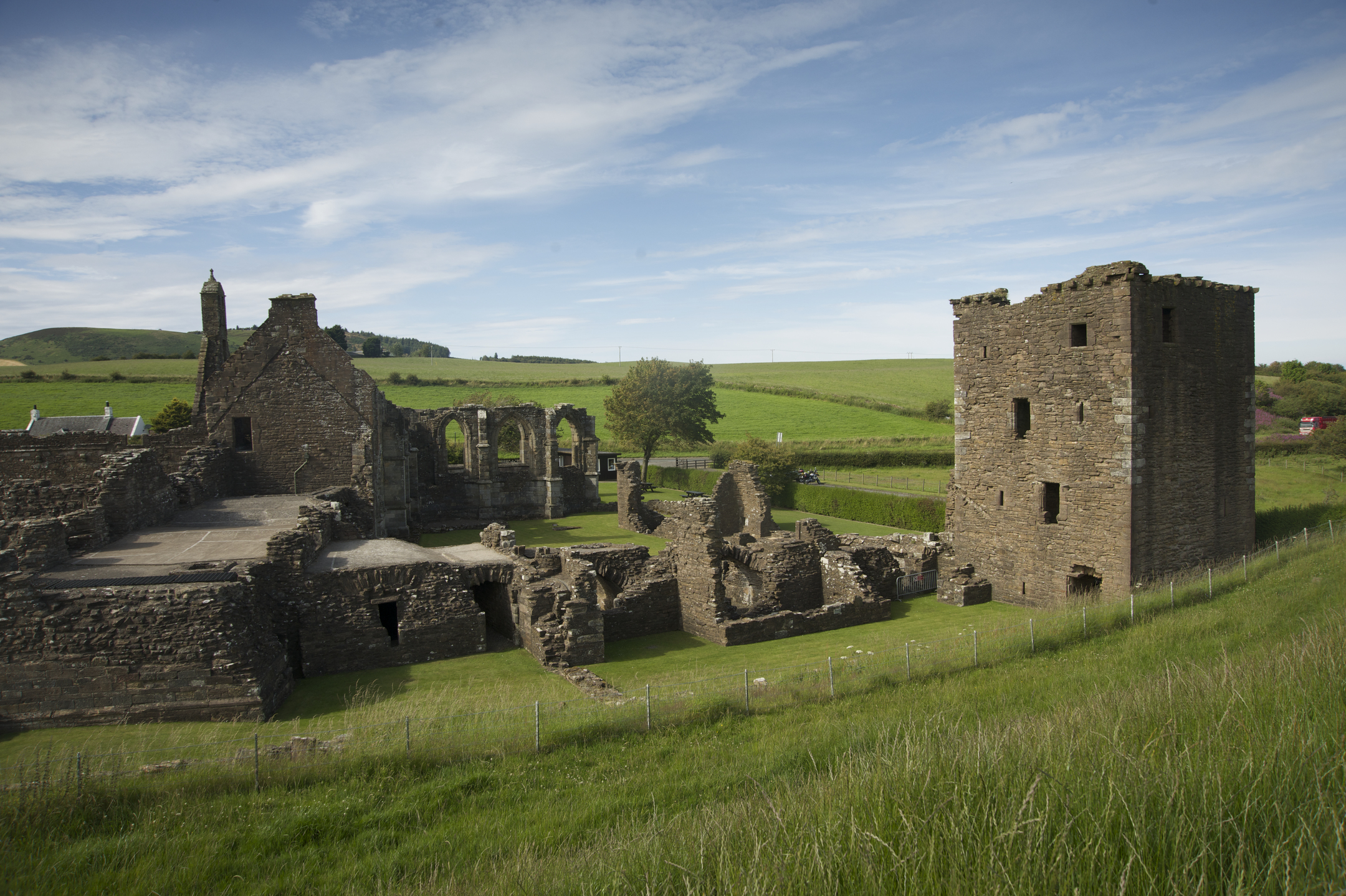 Wide view of the Crossraguel Abbey ruins set in open countryside, with grassy fields and rolling hills behind the remaining stone walls and tower.