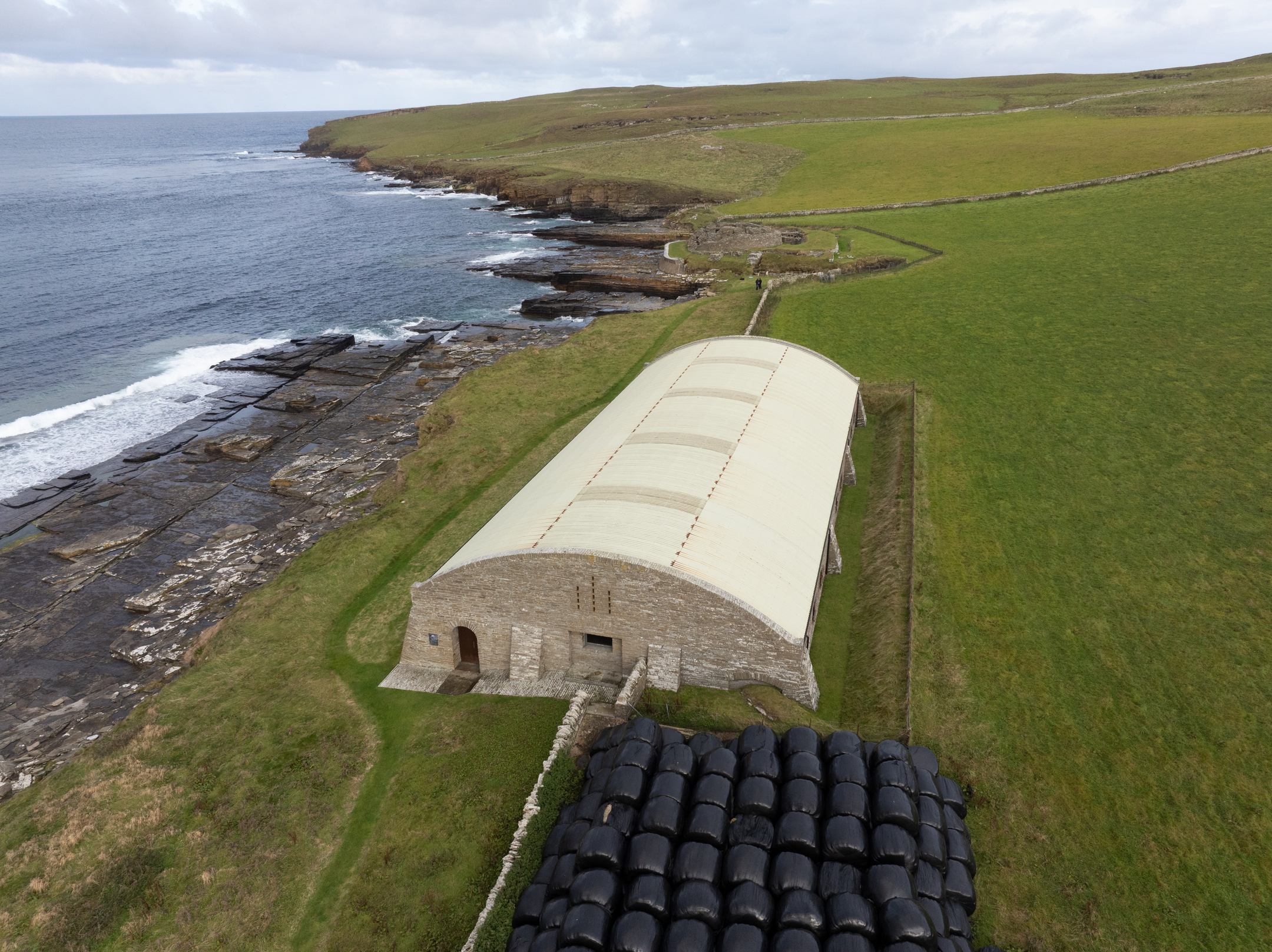Oblique aerial view of the protective hangar beside the shoreline, with cliffs and the Atlantic waves breaking along the rocky coast.