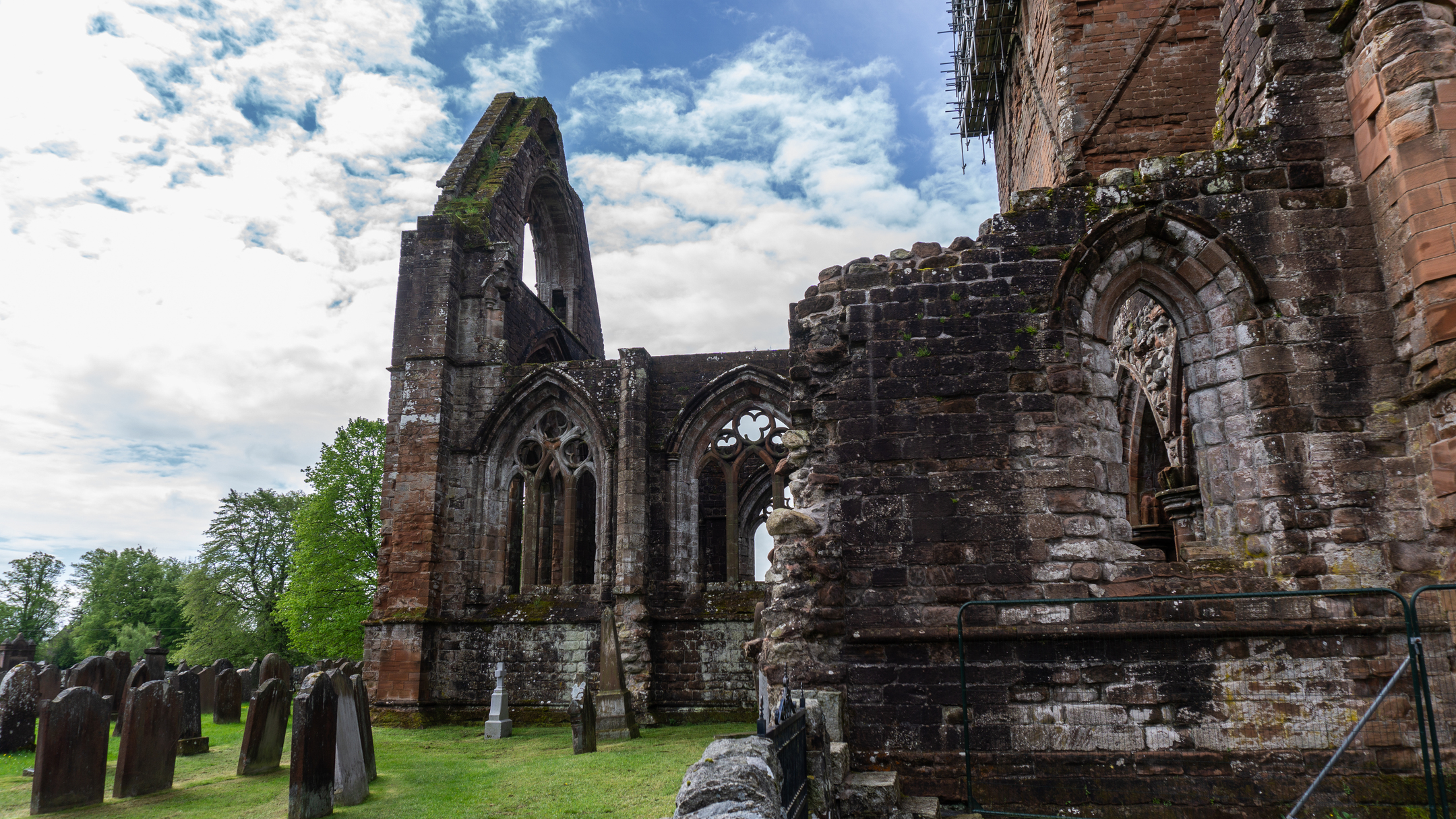 Side of a ruined abbey with prominent window details and gravestones on the left