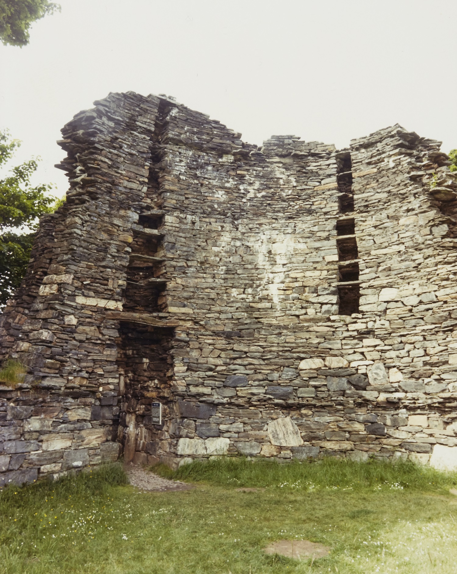 A 1987 colour photograph showing a tall curved section of a broch wall made of stacked stone. Several small openings are visible high up in the thick wall. The structure stands on short grass, with bright sky above and some foliage at the edges of the frame.