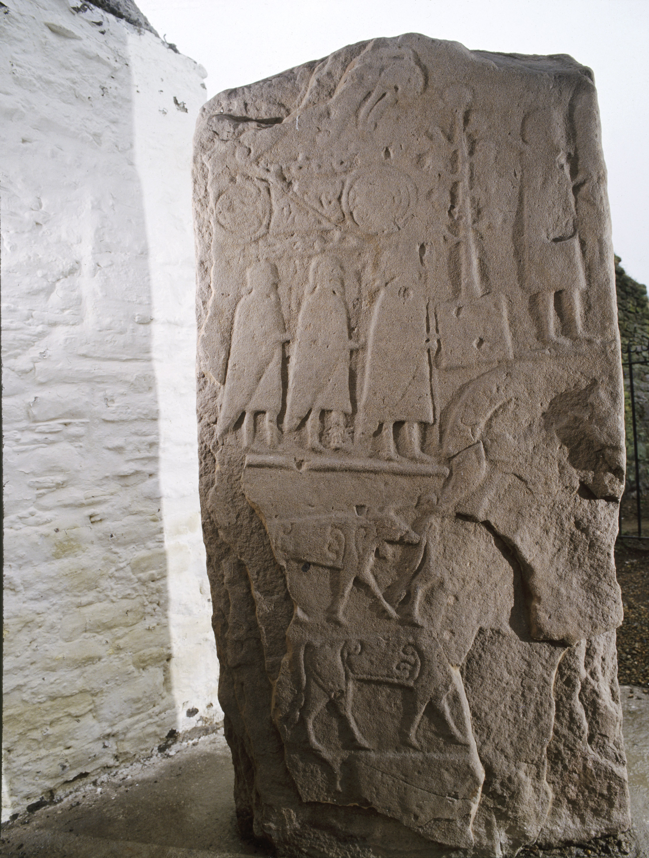 Rear face of the Eassie Pictish sculptured stone, showing worn but recognisable carved figures and symbols. The tall sandstone slab features a row of standing robed figures near the top, with additional human figures and animals carved below, including a person leading an animal and another figure holding a staff or spear. The surface is weathered and uneven, with the carvings illuminated from one side.