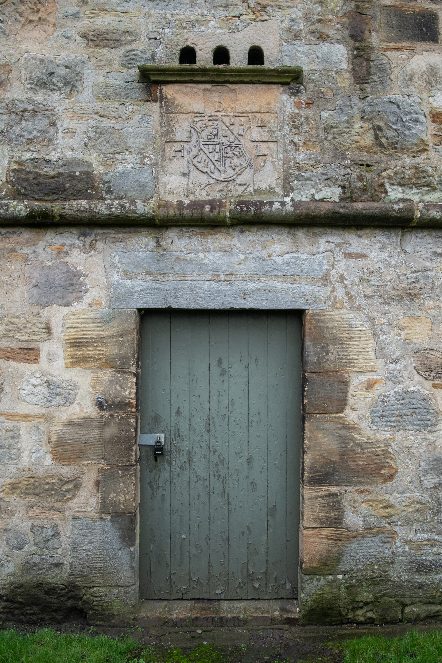 Close-up of the lower façade of the dovecot, showing the green wooden door with metal latch and the carved heraldic panel above it set into the stone wall.