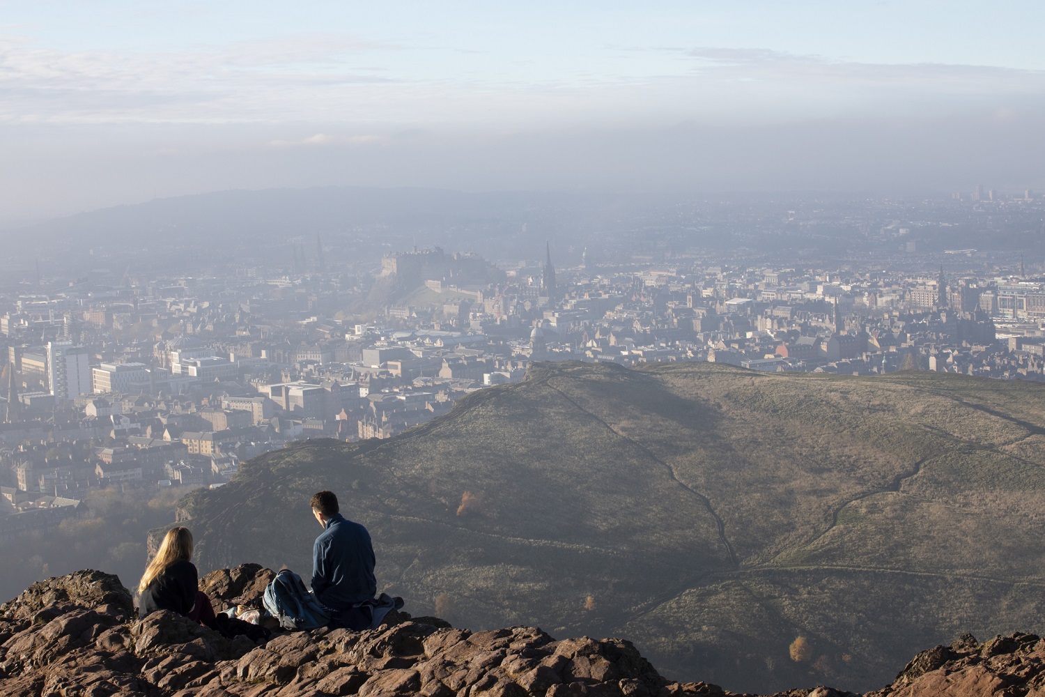 View from Arthur’s Seat showing rocky foreground, two people seated, and Edinburgh cityscape with Edinburgh Castle visible in the distance under a hazy sky.