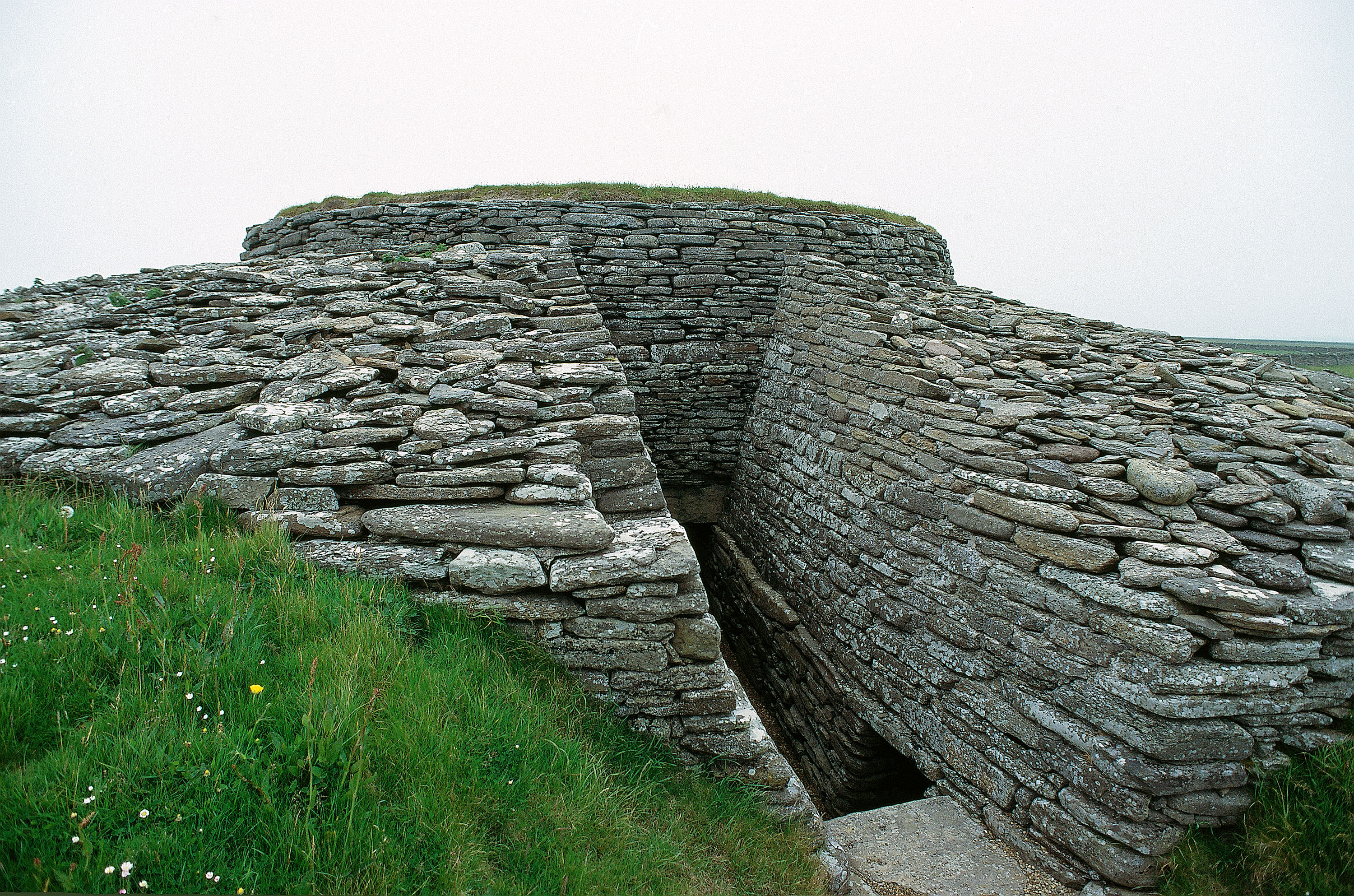 Entrance passage of Quoyness Chambered Cairn, showing a long, narrow, stone‑lined corridor descending between thick drystone walls. The passage cuts into the side of the circular cairn, whose layered stone construction is clearly visible. Grass grows around the outer edge of the mound, and the sky above is overcast, creating a muted, grey light over the scene.