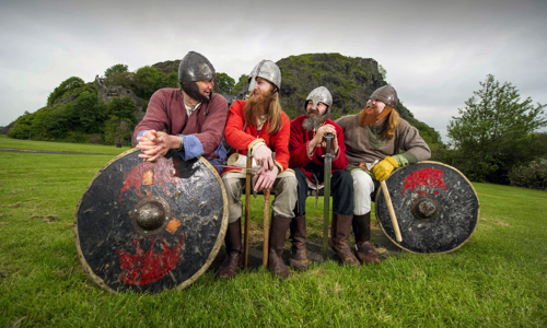 Four men in Viking costumes with helmets on are sitting on a bench looking at each other surrounded by grass. The two Vikings on the outside have shields in front of them while the two in the middle have swords. Dumbarton Rock is in the background.