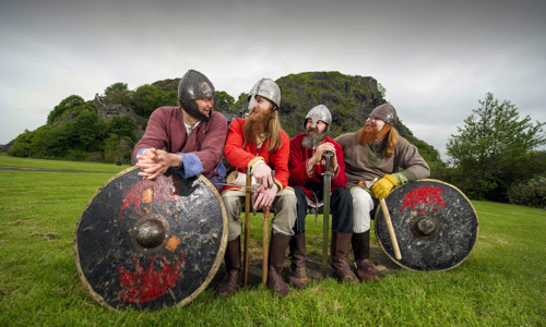 Four men in Viking costumes with helmets on are sitting on a bench looking at each other surrounded by grass. The two Vikings on the outside have shields in front of them while the two in the middle have swords. Dumbarton Rock is in the background.
