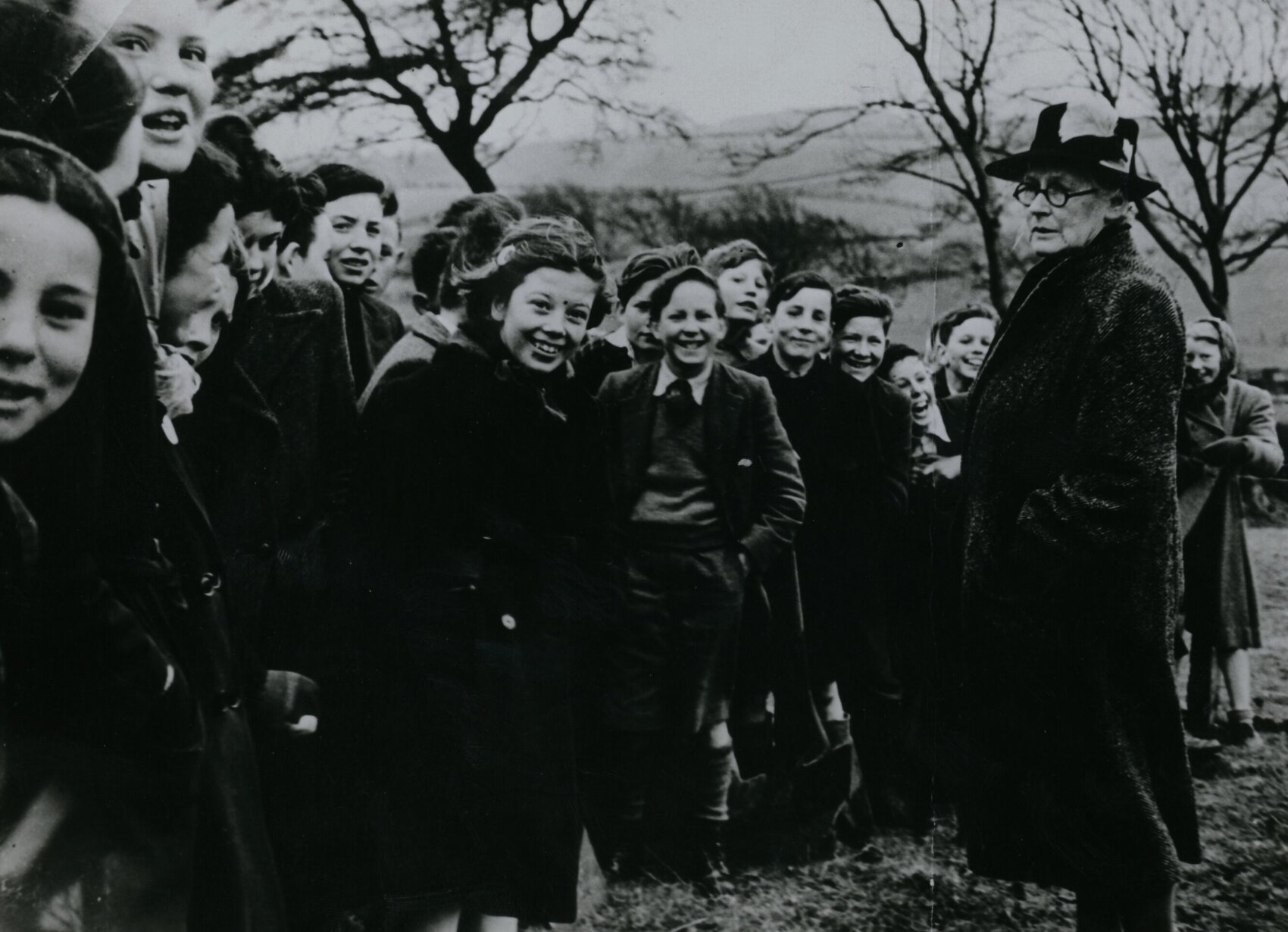 Black and white photo of a group of 1950s schoolchildren and a person supervising them. They are at a tree planting ceremony, and the uniformed children are laughing and smiling. The person is looking sternly at them, with their hands in their coat pockets.