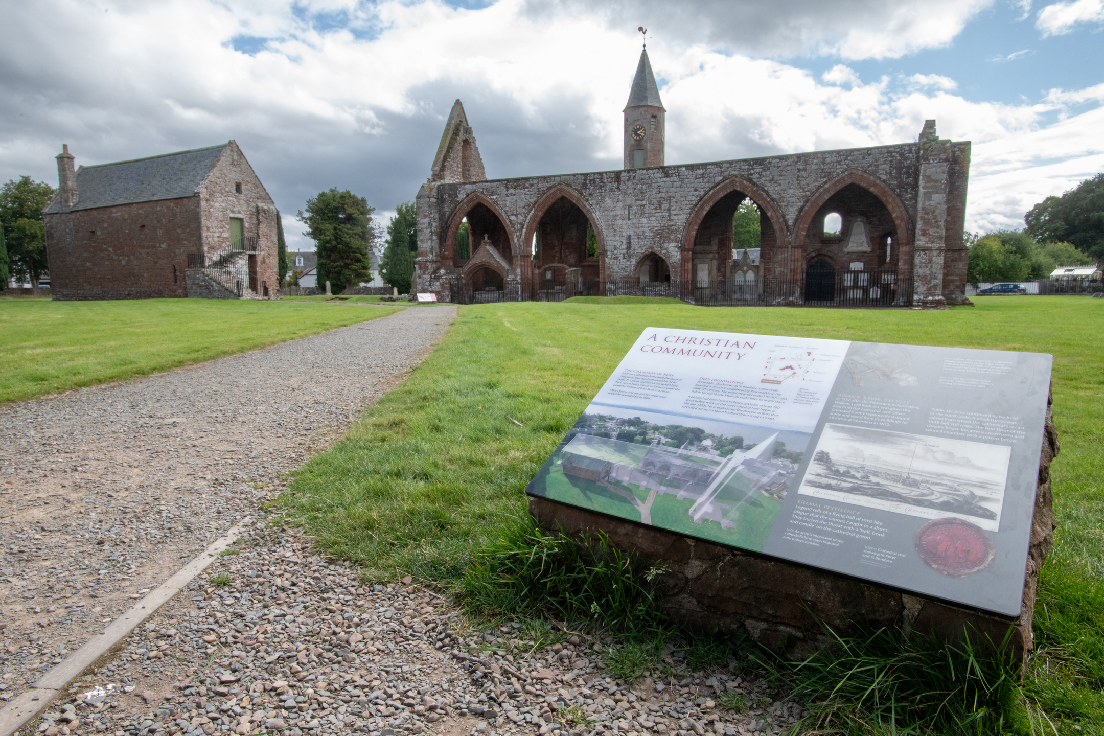 Wide view of Fortrose Cathedral ruins seen across a grassy field, with a large outdoor interpretive panel in the foreground titled ‘A Christian Community,’ showing illustrations, maps, and historical information about the cathedral’s layout and history