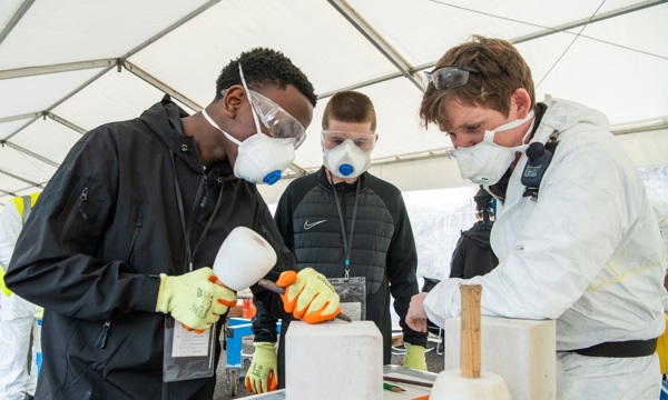 Three individuals wearing protective gear - gloves, goggles, and face masks - work together under a white tent, carving stone blocks using chisels and mallets. The scene captures a hands-on training workshop teaching traditional stonemasonry skills.