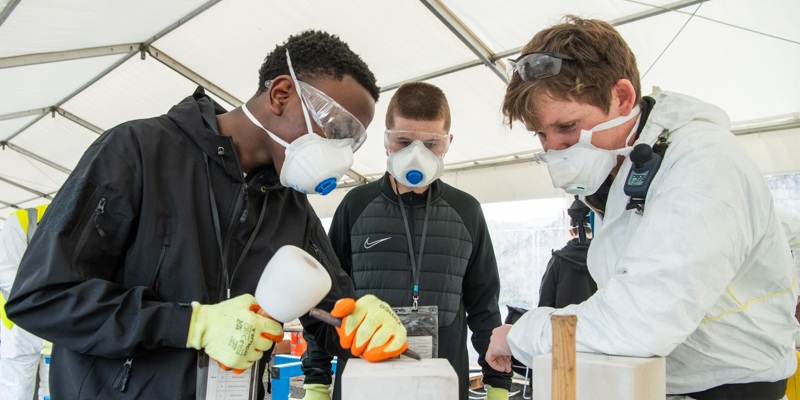 Three individuals wearing protective gear - gloves, goggles, and face masks - work together under a white tent, carving stone blocks using chisels and mallets. The scene captures a hands-on training workshop teaching traditional stonemasonry skills.