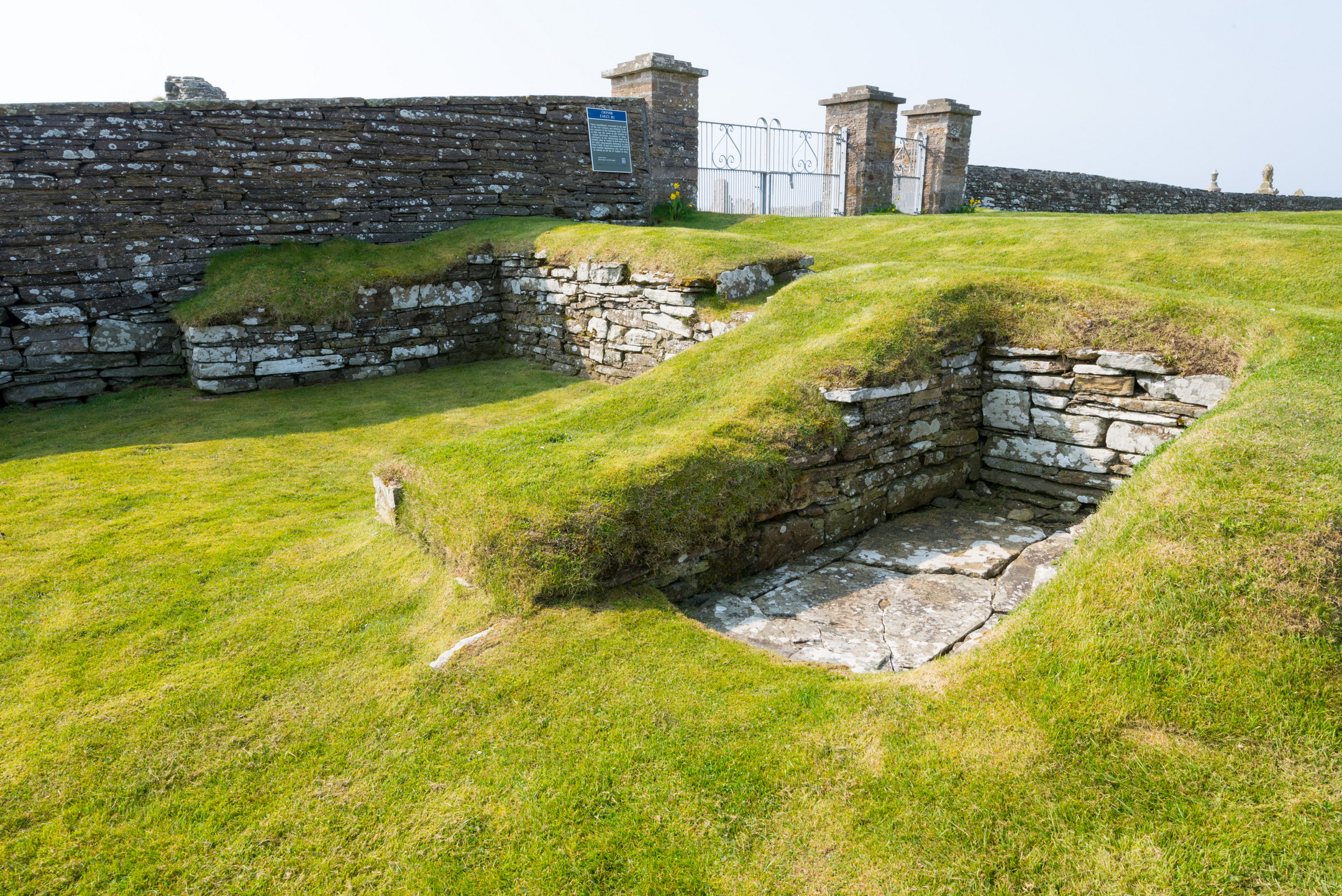 Grassy area with wall and gate in background and remains of building in foreground
