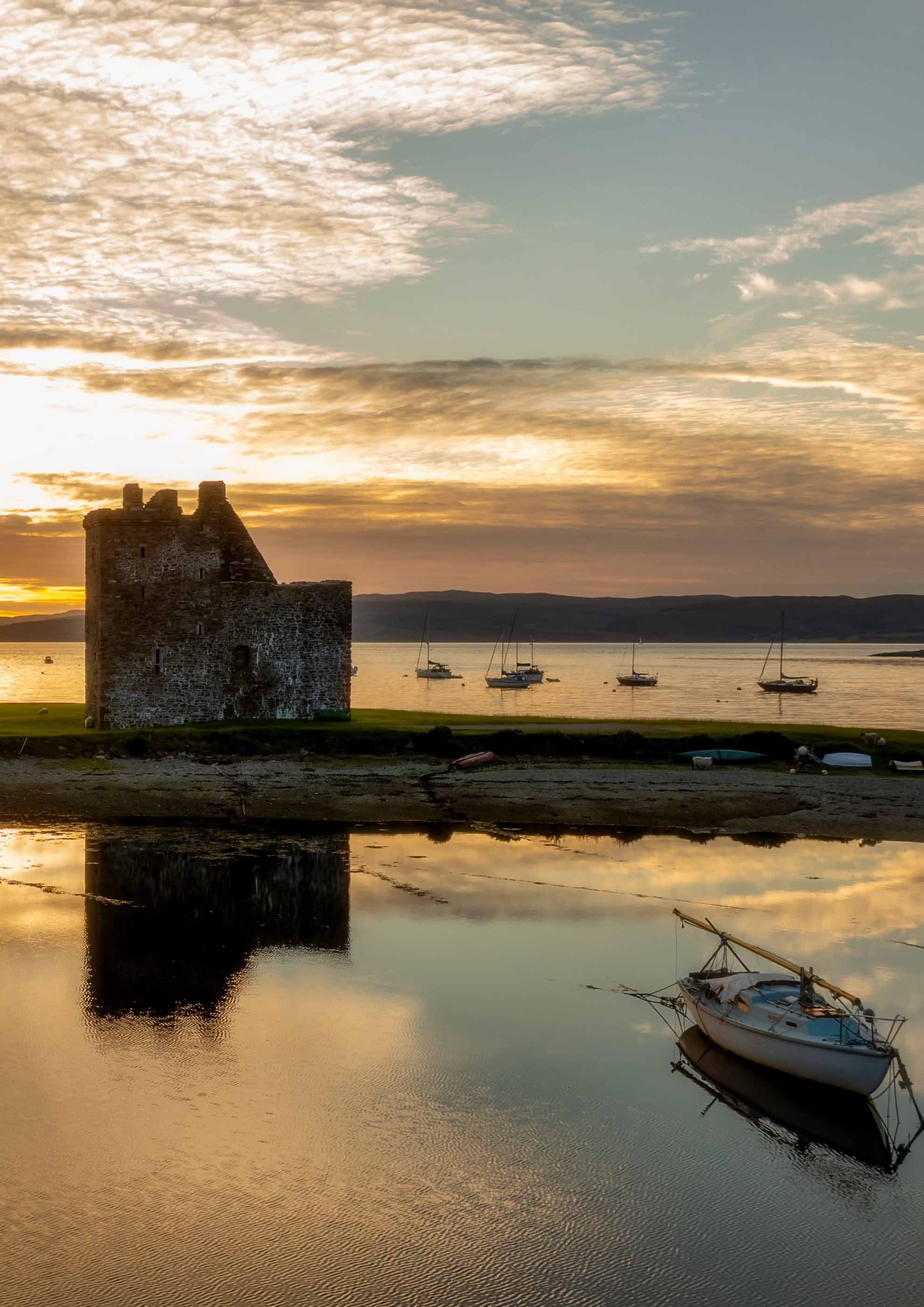 A stone castle on a narrow strip of land surrounded by the sea at sunset, with a boat in front and a row of boats behind it