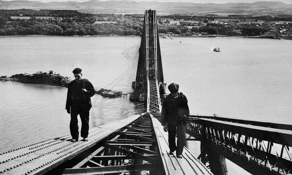 A black and white photo of two people walking along the edge of a metal bridge. More of the bridge is visible behind them, with the water of the Firth of Forth below and land in the distance.
