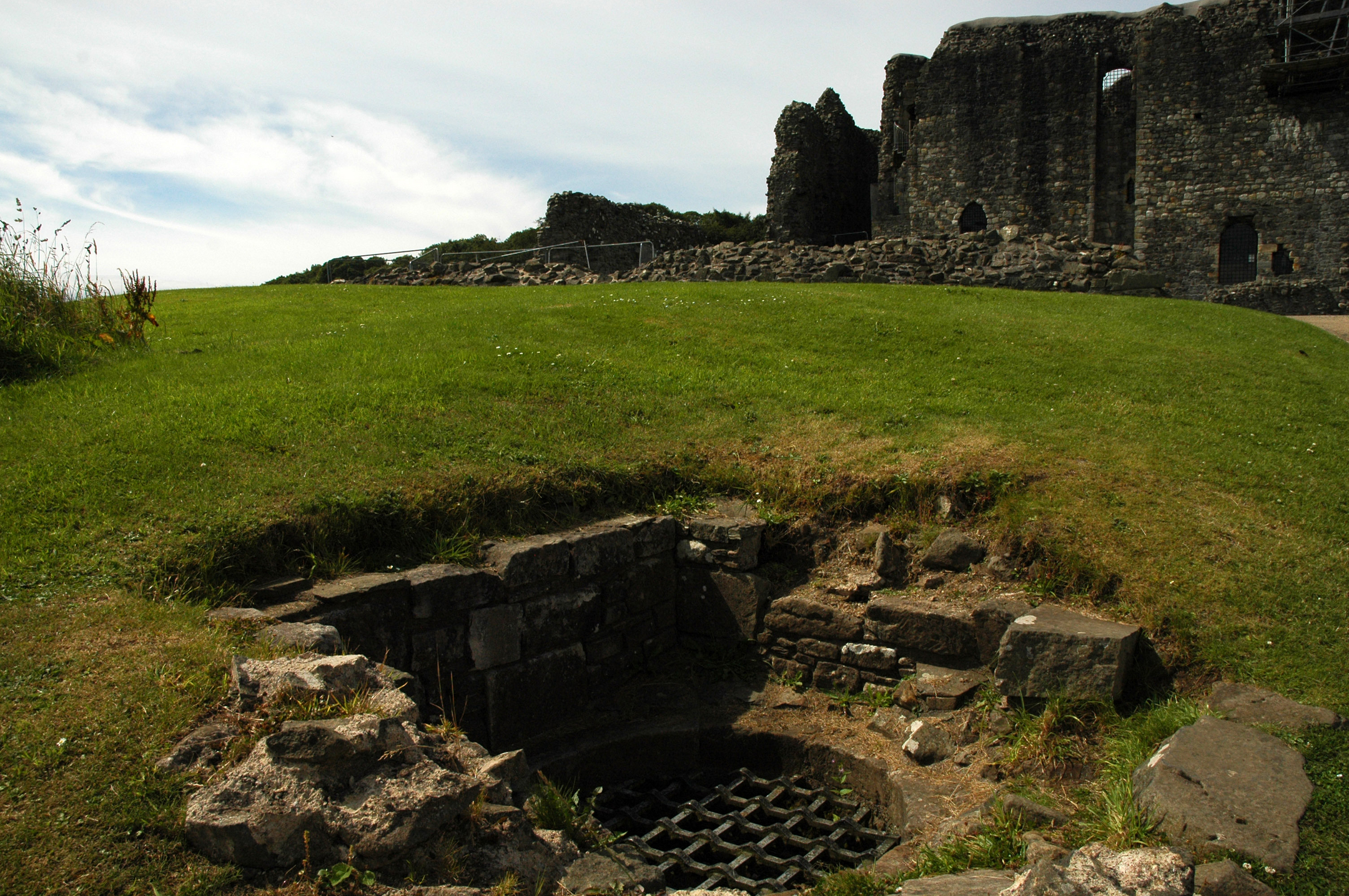 A small stone-lined well in the foreground, with the castle ruins rising on the hill behind it.