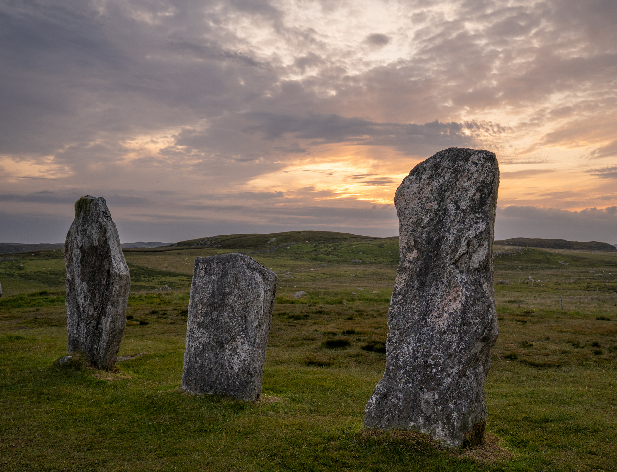 Three Calanais stones in a row with a gentle, glowing sunset behind them.
