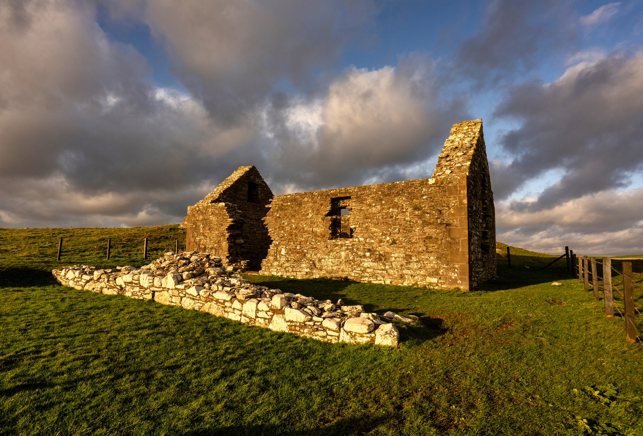 Close view of St Ninian’s Chapel ruin and a low stone foundation beside it, illuminated by golden-hour sunlight on a grassy slope.
