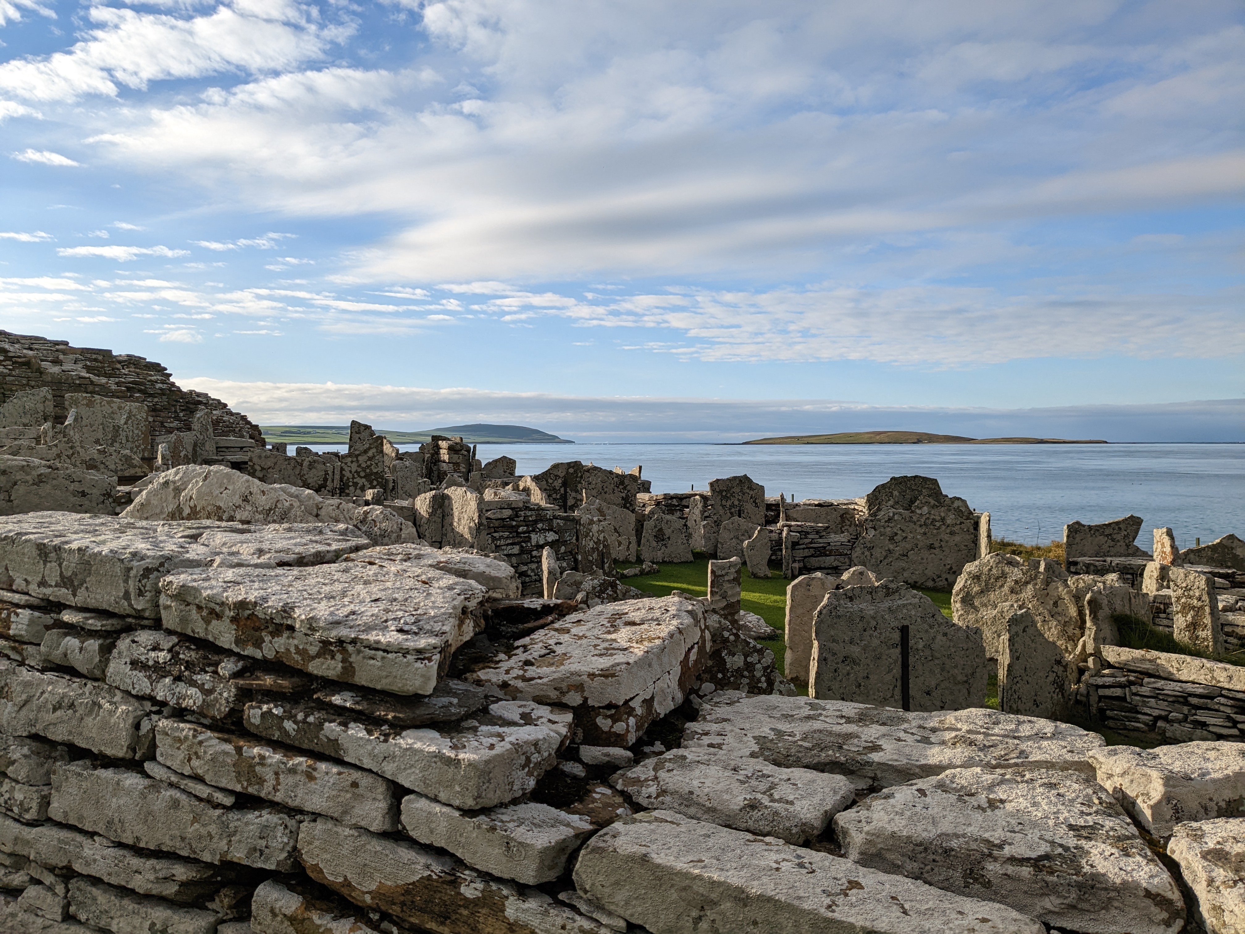 Stone ruins of the Broch of Gurness with upright slabs and layered walls, set against a calm blue sea and distant islands under a partly cloudy sky.