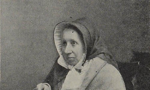 Sepia toned, older newspaper image of woman sat for portrait photo facing at a 45 degree angle. She is wearing period clothing, appears to be wrapped in a shawl and holds her round metal glasses in her right hand.