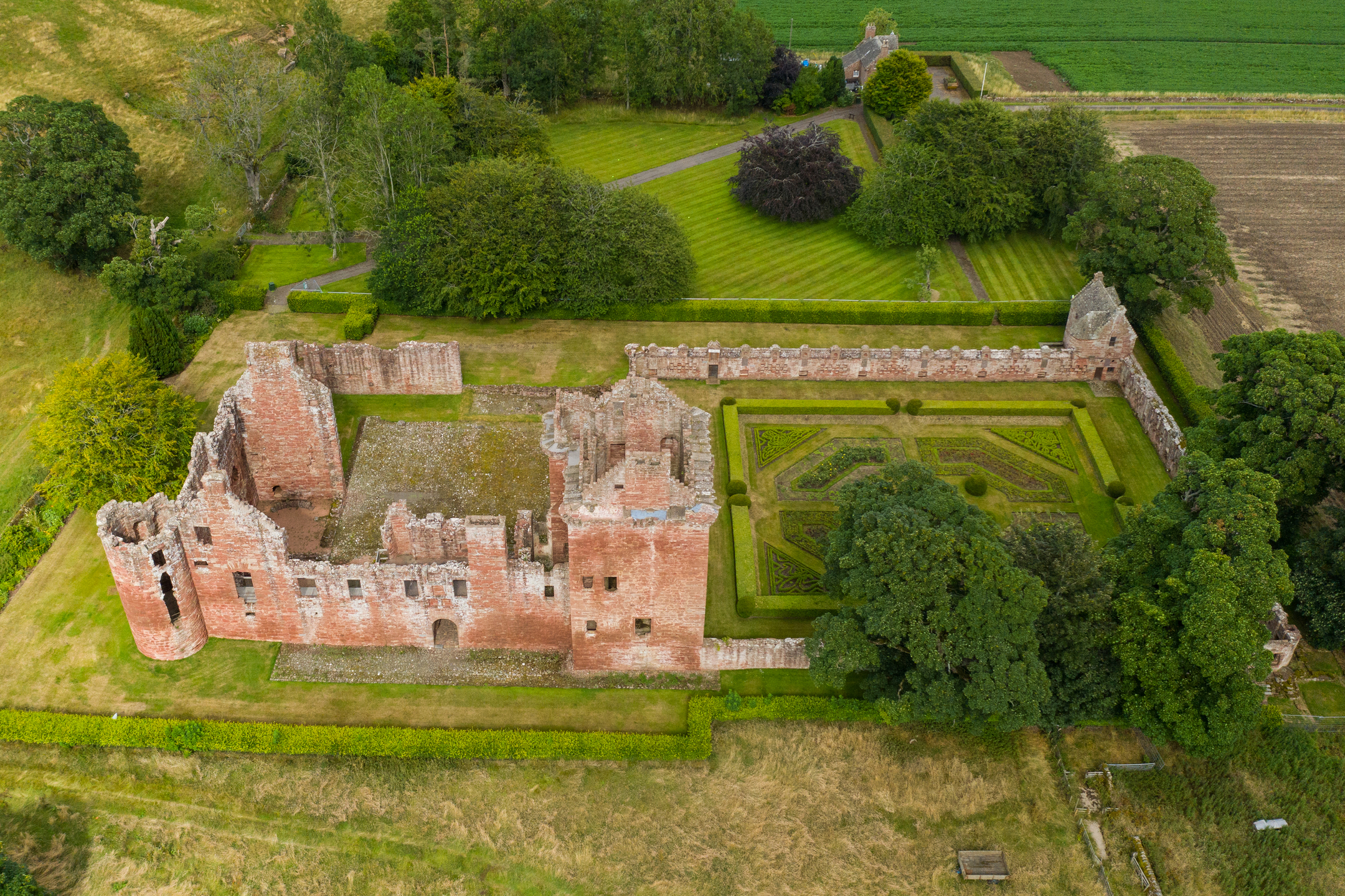 An aerial photo of a red sandstone castle beside an ornate walled garden. The tower house in well-preserved, while the remaining rooms, walls and towers are in more ruinous state. A pathway leads across green parkland from a small visitor centre.