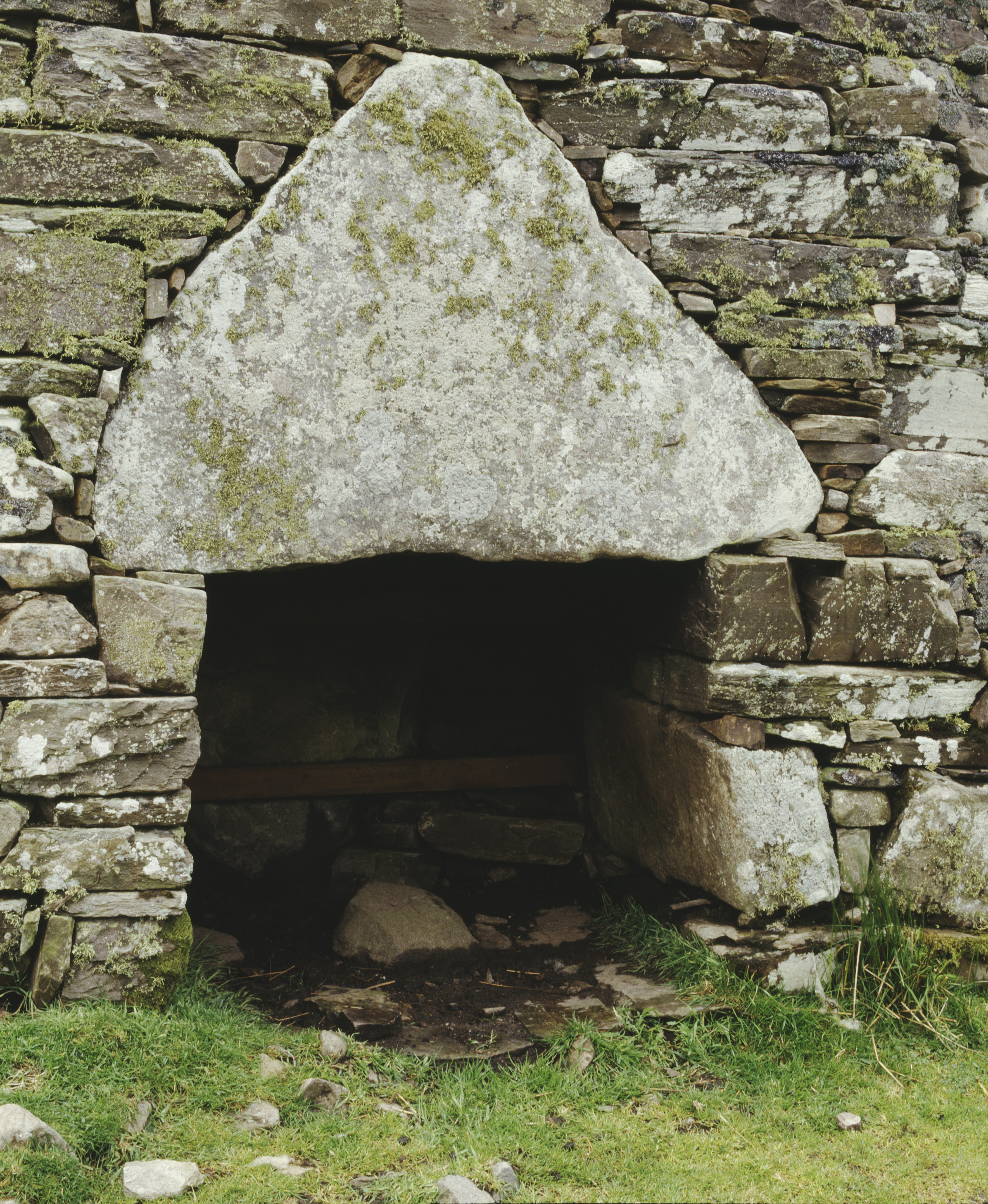 Close-up view of the entrance to Dun Dornaigil broch, showing a narrow doorway framed by large, weathered stone blocks. A large triangular lintel stone sits above the opening, covered in patches of lichen. The stones around the entrance are irregular and tightly fitted, with grass and small rocks at the base.
