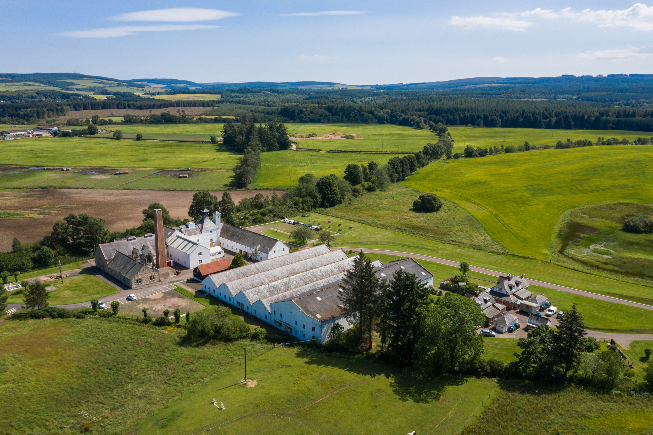 Aerial view of Dallas Dhu distillery set within rolling green countryside, with white industrial buildings, fields, woodland, and distant hills.