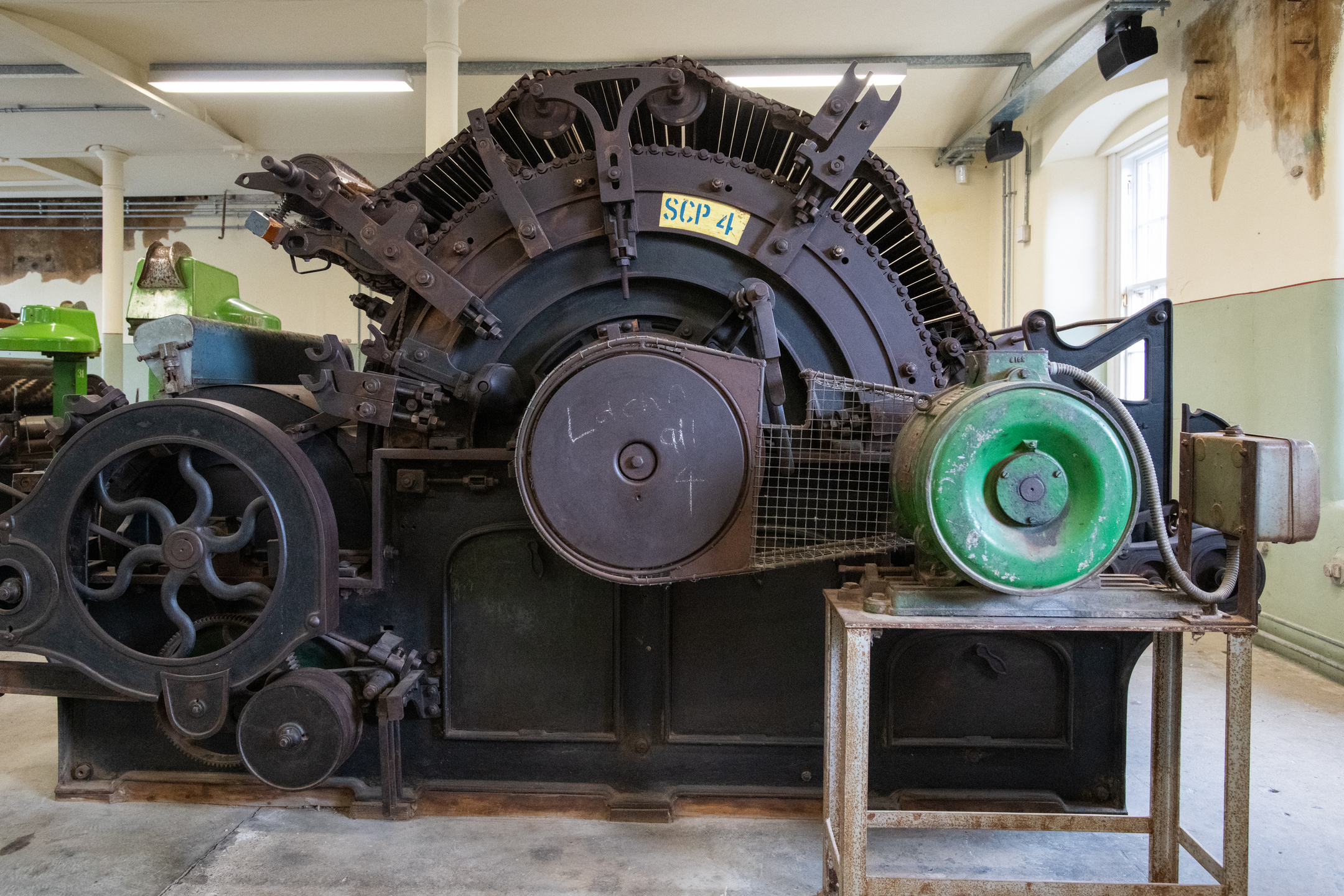 A large, heavy industrial spinning or carding machine with gears, rollers, and a green motor sits inside the mill’s machinery hall, showing rust and wear.