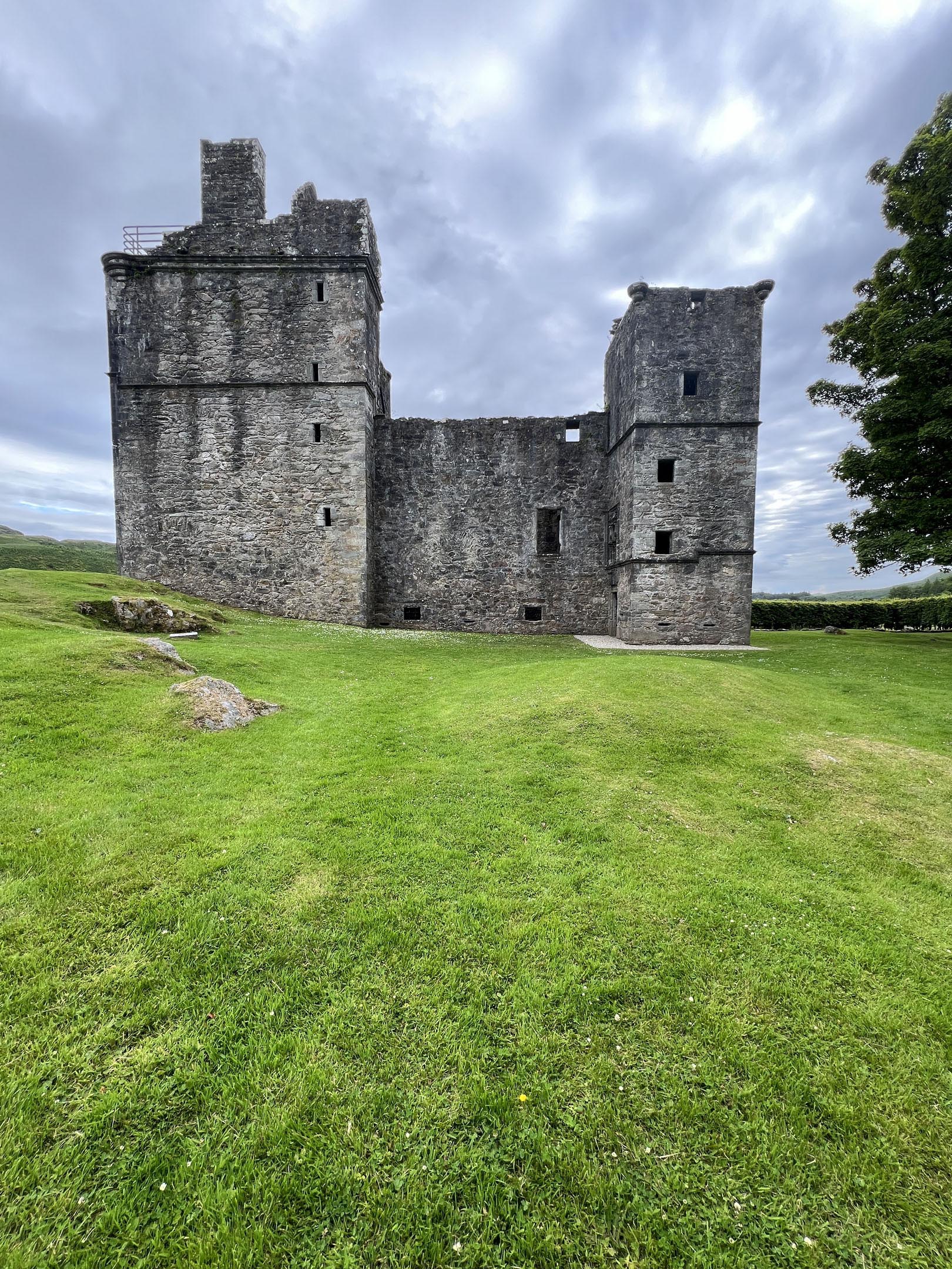Stone castle ruins with two towers on grassy slope under cloudy sky.