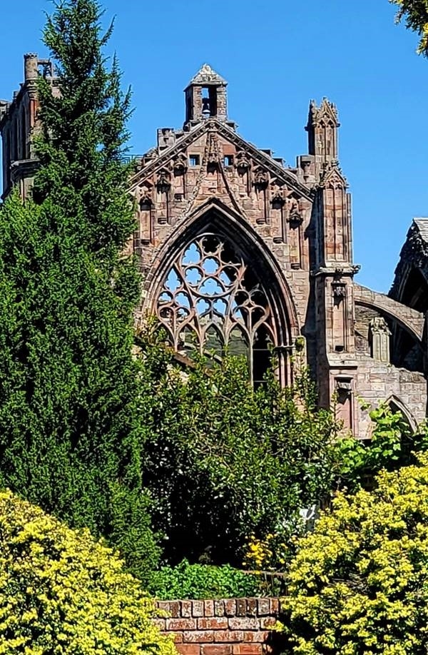 Large trees and bushes stand in front of part of a historical building, which is in the shape of an arch. There is detailing on the arch and you can see through the pttern. There are blue skies in the background. 